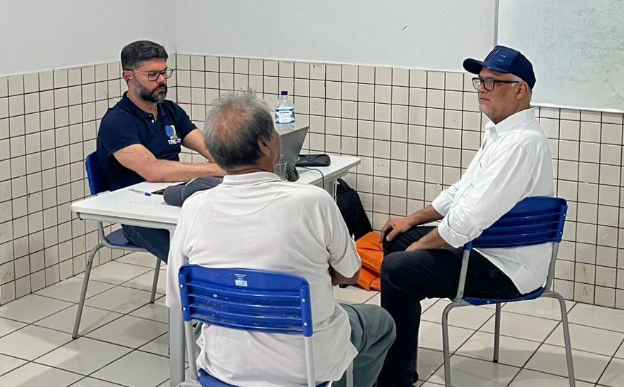 O juiz Wellington Magalhães realiza atendimento em sala, sentado à mesa com equipamento de trabalho, enquanto dois homens são atendidos, em ambiente adaptado para a ação itinerante.