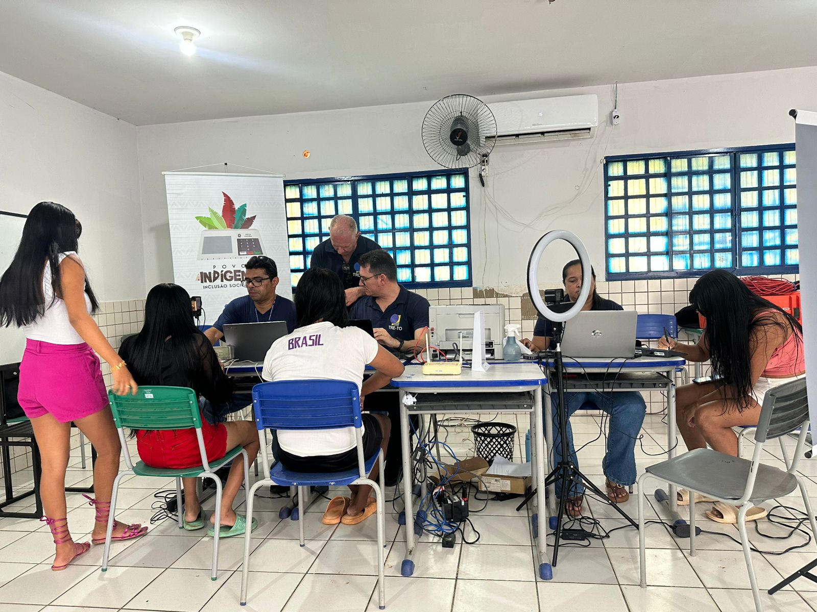 Atendimento itinerante em sala adaptada, com servidores utilizando computadores para prestar serviços à população indígena. Pessoas aguardam e são atendidas em mesas organizadas, com uso de equipamentos como ring light para registro. Banner ao fundo indica ação voltada à inclusão social dos povos indígenas.
