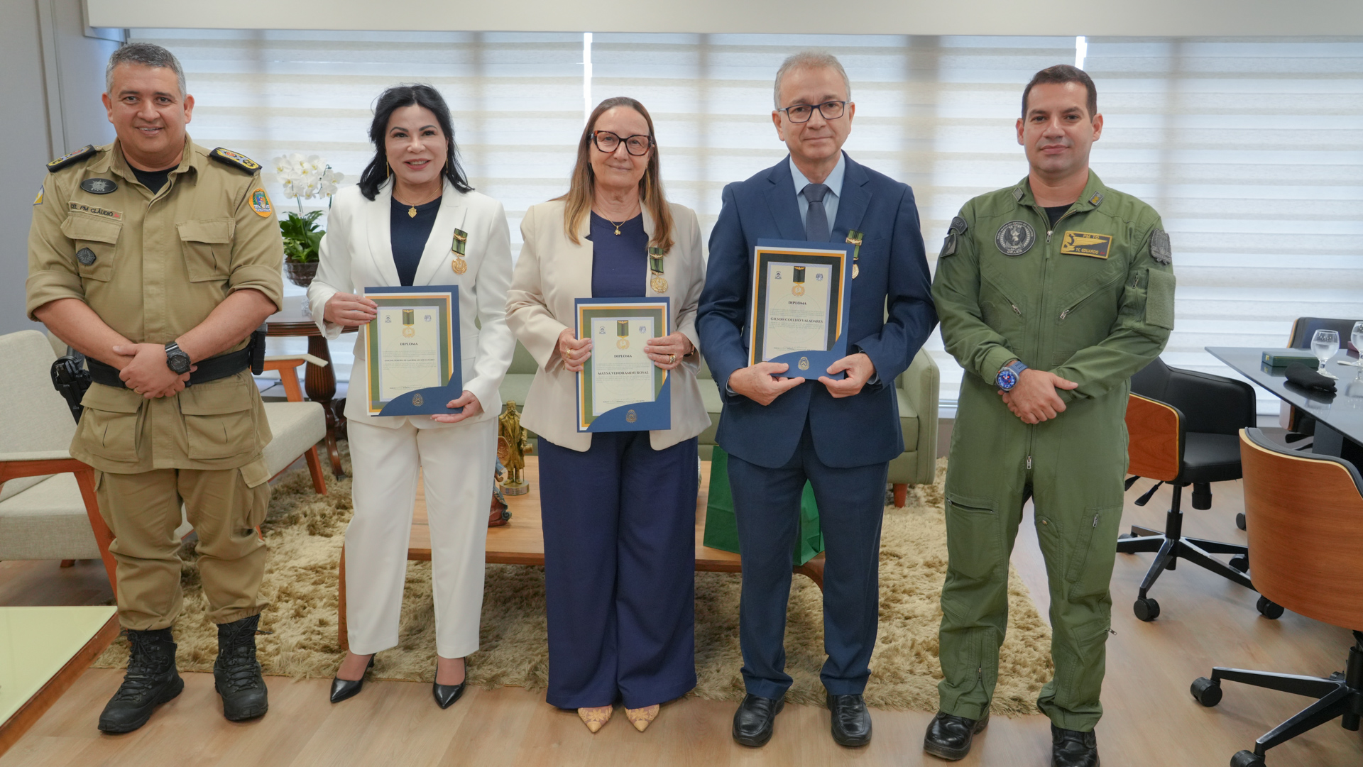 Cinco autoridades posam em gabinete institucional. Três desembargadores, ao centro, seguram certificados e usam medalhas. À esquerda, comandante da PM em uniforme cáqui; à direita, comandante em traje de aviação. Ambiente formal, com mobiliário e cortinas claras ao fundo.