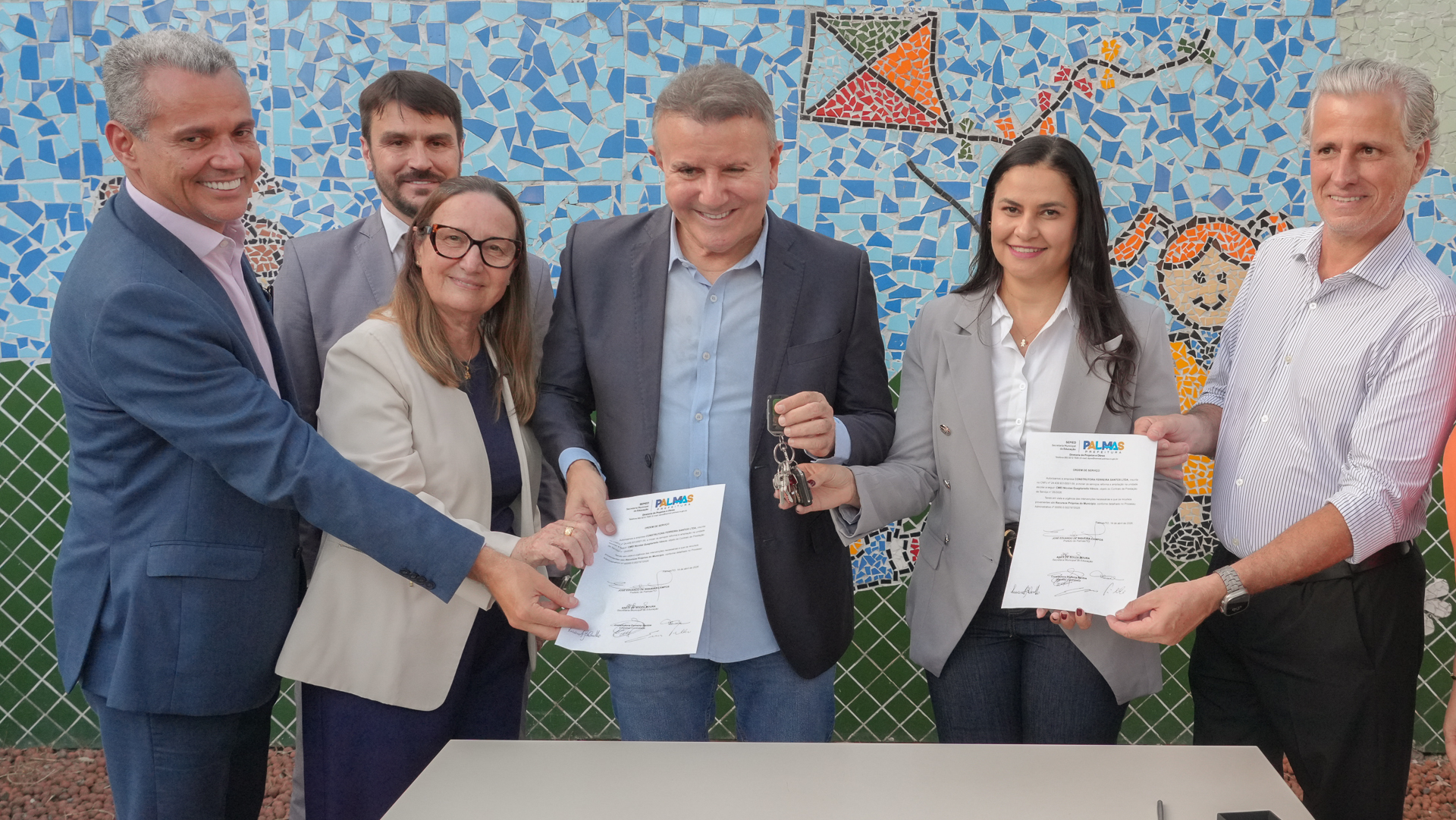 Grupo de autoridades posa para foto durante entrega institucional, segurando documentos e uma chave simbólica. Ao fundo, um mural colorido em mosaico