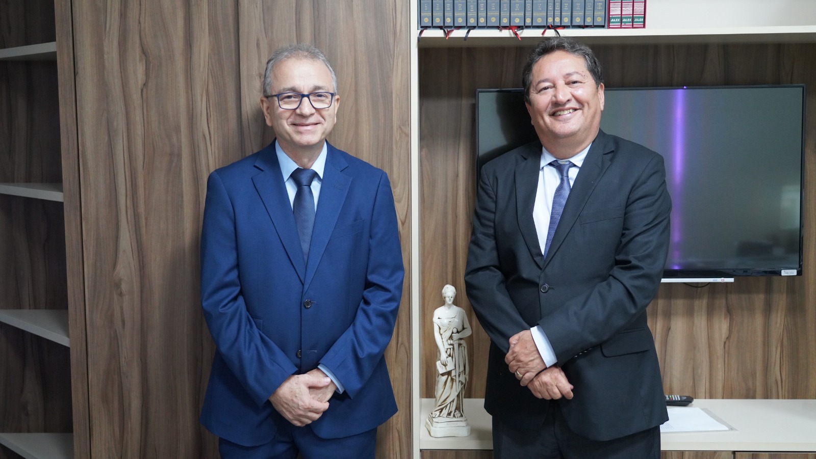 Dois desembargadores posam lado a lado em gabinete institucional, vestindo ternos escuros e gravatas. Ao fundo, estante com livros jurídicos, uma televisão e uma escultura decorativa, compondo ambiente formal e organizado.