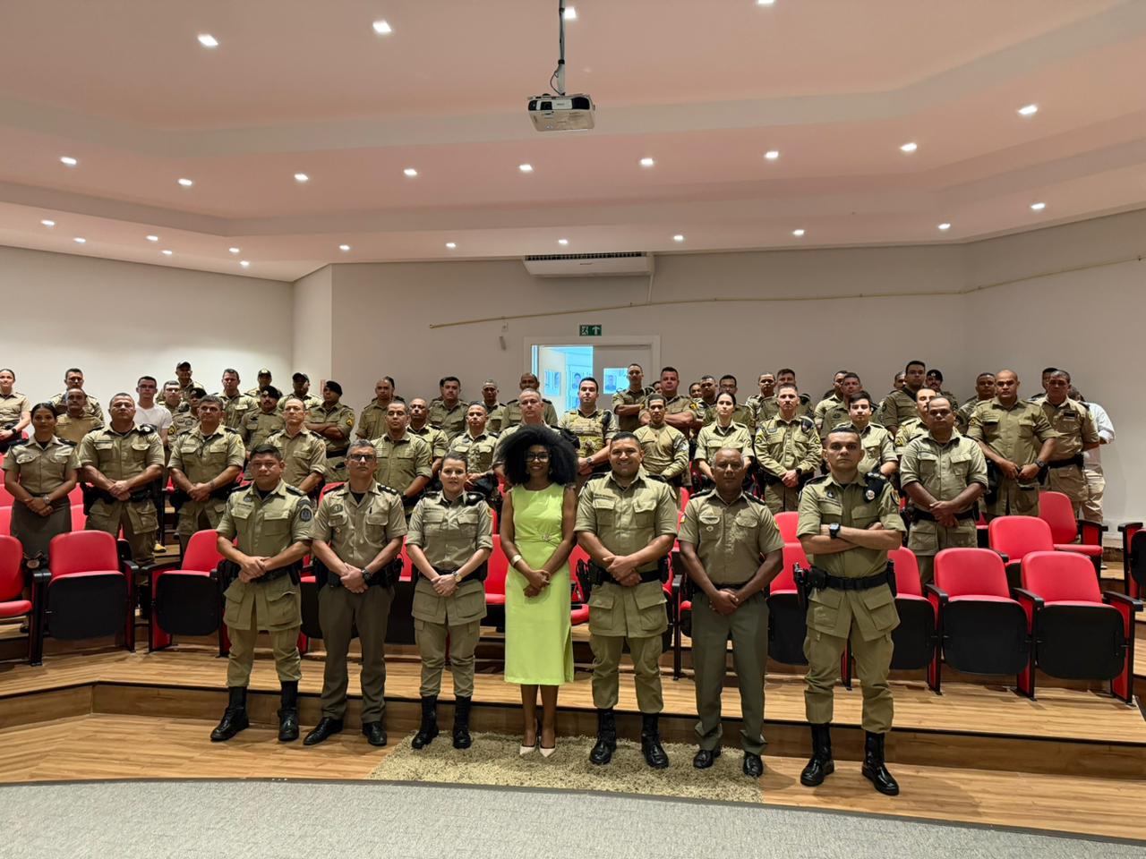Auditório com a juíza e dezenas de policiais militares fardados, organizados em fileiras. Na parte frontal, um grupo posa para a foto, com destaque para a juíza ao centro, usando vestido amarelo, cercada por integrantes da corporação. O ambiente é fechado, com cadeiras vermelhas, iluminação embutida no teto e projetor instalado acima.