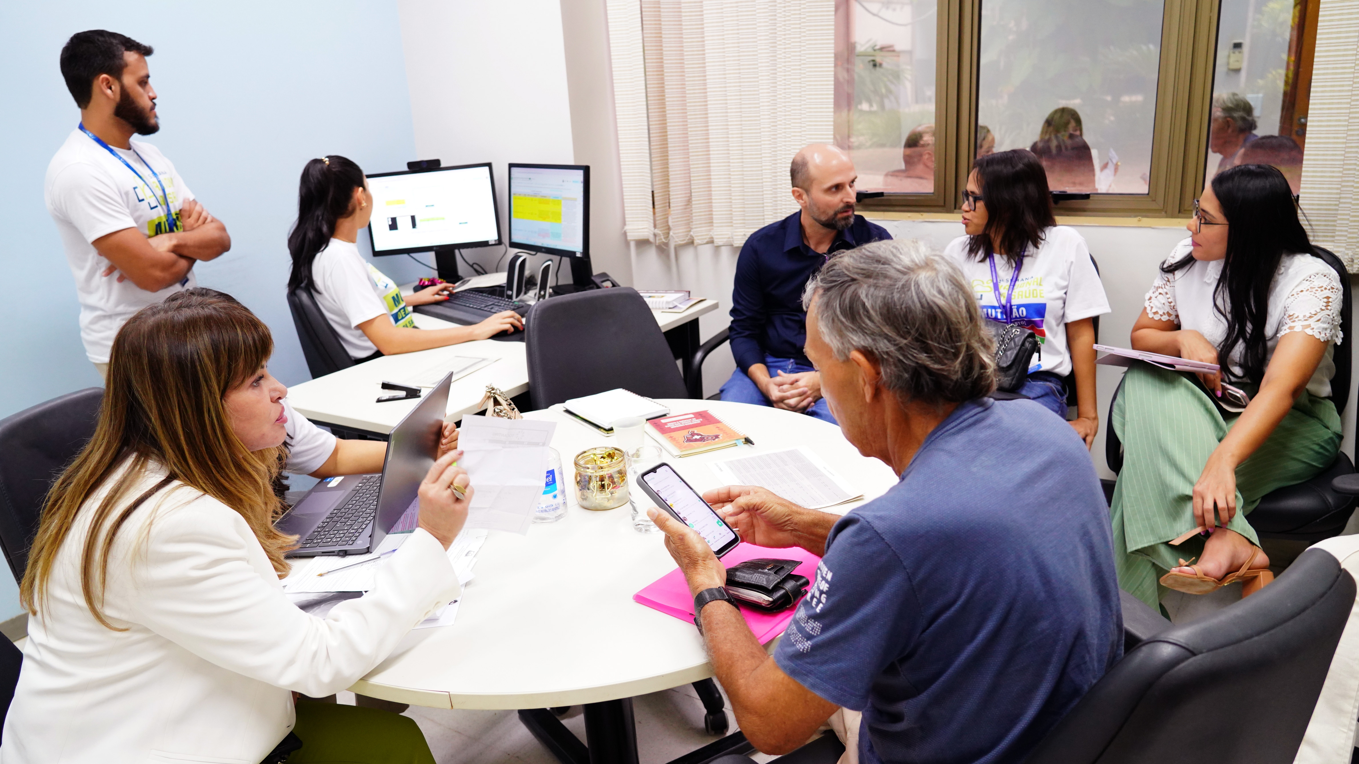 Atendimento de conciliação em saúde reúne equipe técnica e usuários em sala do Judiciário. Profissionais analisam documentos e sistemas, enquanto um paciente é orientado durante o acompanhamento do caso.