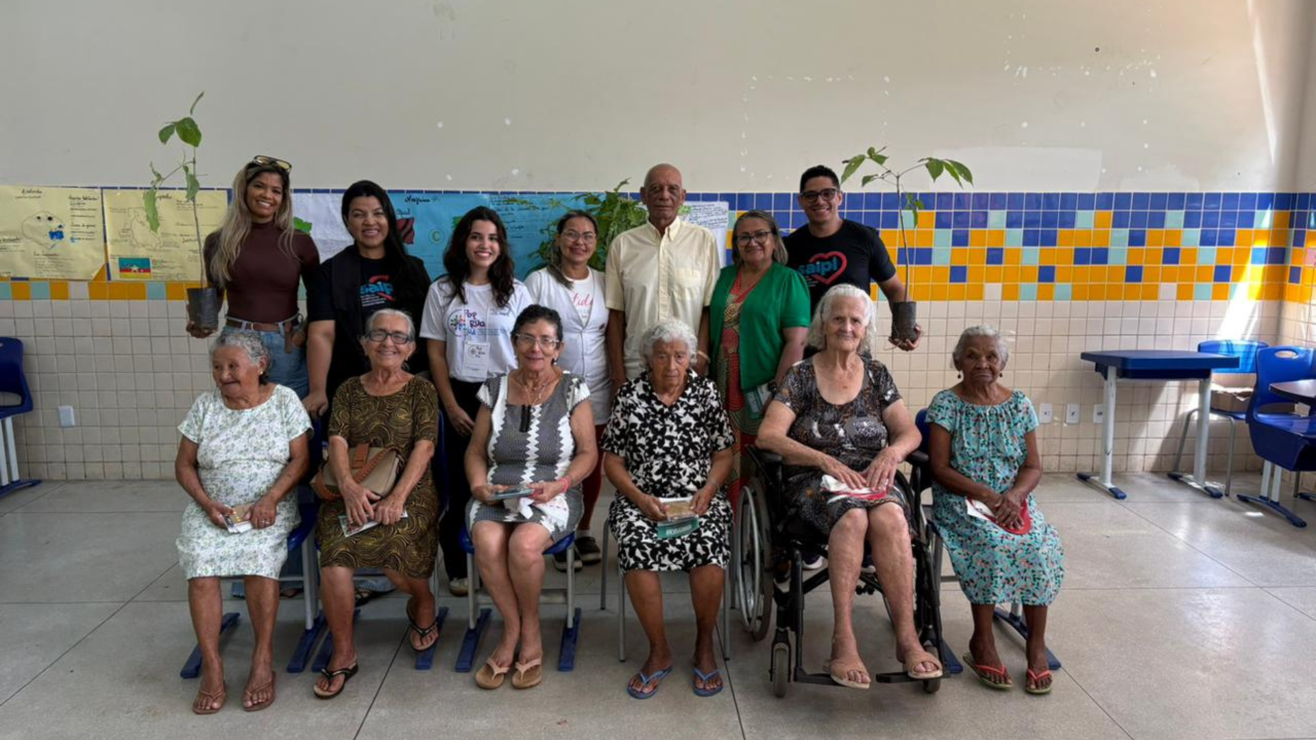 A imagem mostra um grupo de pessoas de diferentes idades reunidas em uma sala, com destaque para idosas sentadas na frente e outras pessoas em pé atrás, todas sorrindo e segurando materiais impressos.