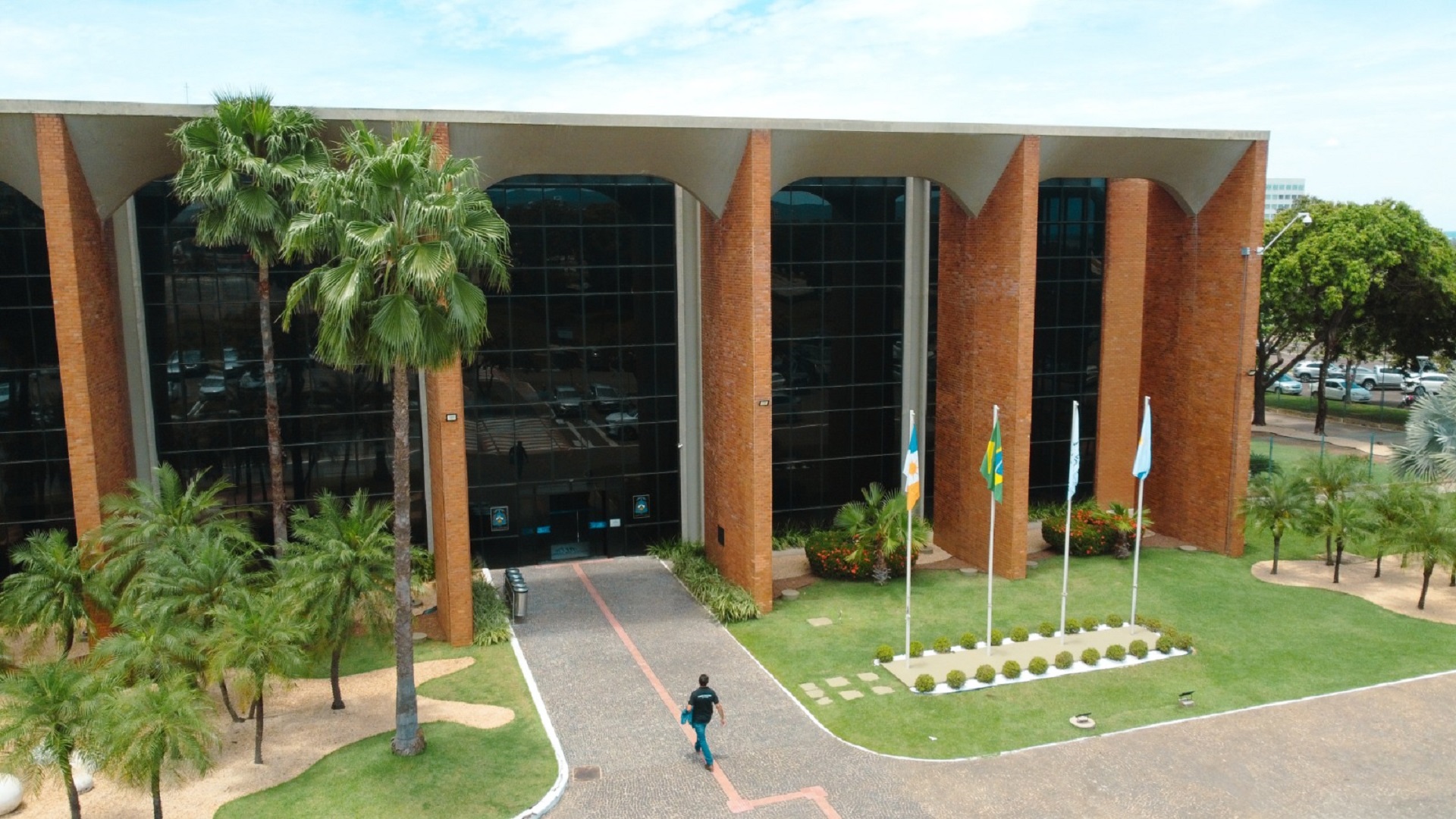 A imagem mostra a fachada do prédio do Tribunal de Justiça do Tocantins, com uma  estrutura em tijolos aparentes, grandes painéis de vidro e palmeiras ao redor, destacando um ambiente bem cuidado e arborizado. Em frente ao edifício, bandeiras estão hasteadas e uma pessoa caminha pela entrada principal.
