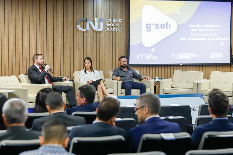 Harley Carreiro, desenvolvedor da Diretoria de Tecnologia da Informação do TJTO, aparece sentado à esquerda do palco durante painel no CNJ. Com microfone em mãos, ele apresenta a ferramenta Giseli enquanto, ao fundo, a projeção exibe a identidade visual da solução. Ao lado, outros participantes acompanham o debate, diante de uma plateia atenta à discussão sobre inteligência artificial no Judiciário.