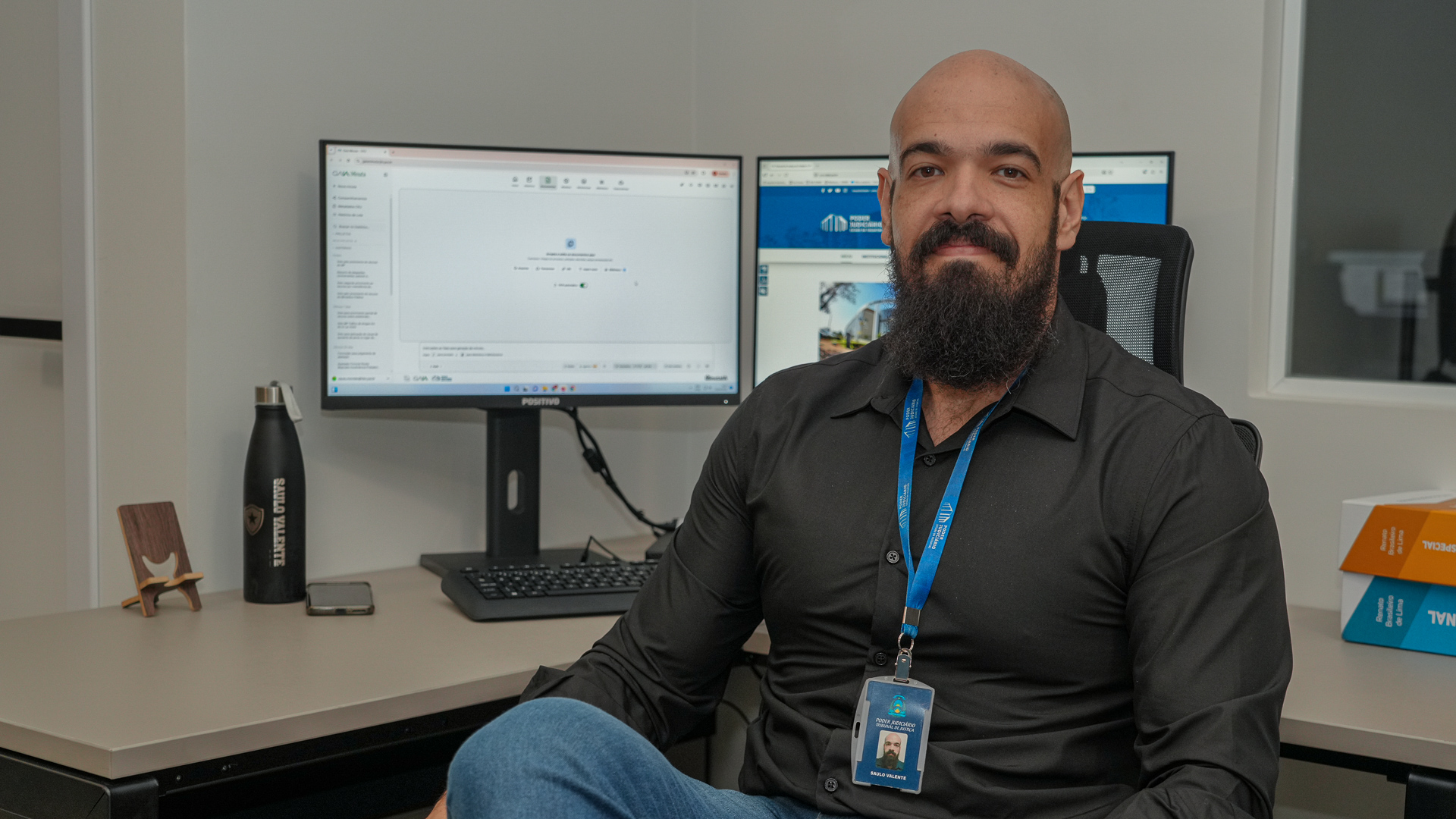 Servidor do Tribunal de Justiça do Tocantins posa em ambiente de escritório, sentado diante de uma estação com dois monitores. Ele veste camisa social preta e utiliza crachá institucional. Ao fundo, uma das telas exibe o sistema GAIA Minuta e a outra o portal do Judiciário. Sobre a mesa, há itens de uso pessoal e de trabalho, como garrafa, celular e suporte, compondo um ambiente organizado e profissional.