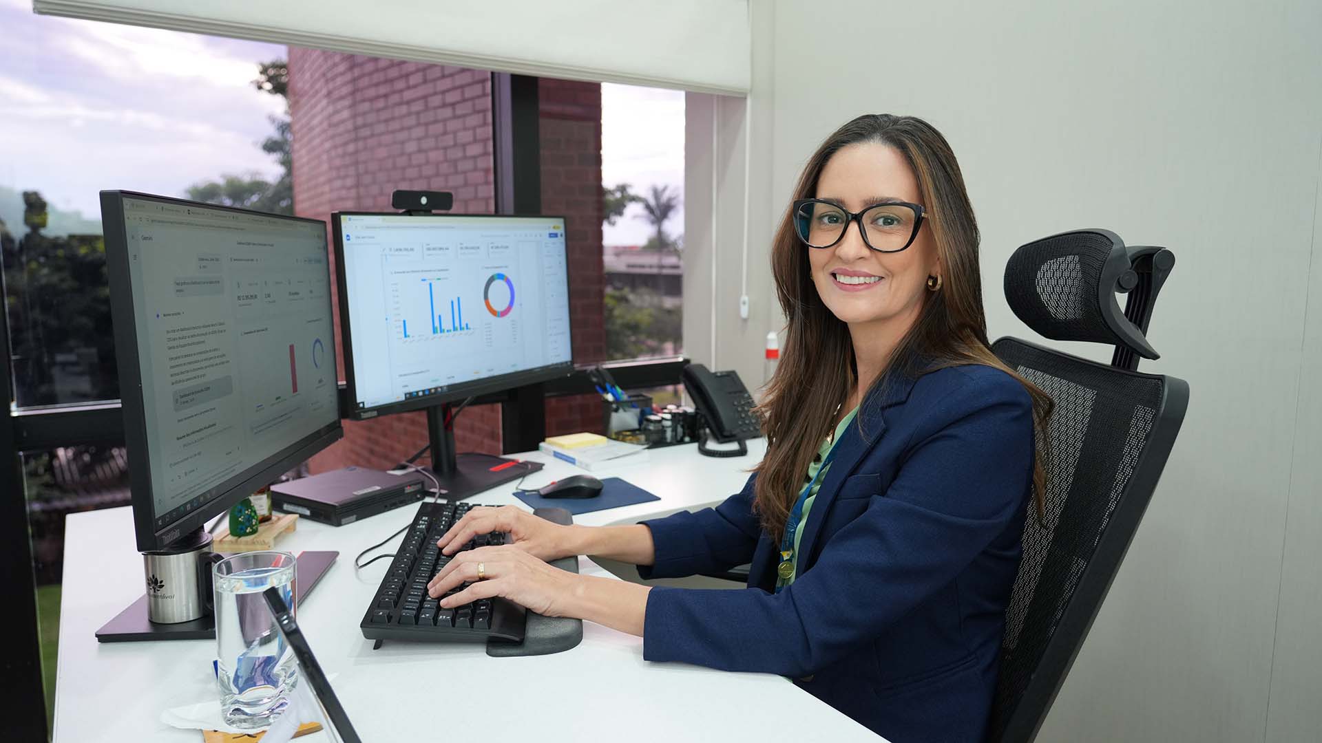 A imagem mostra uma mulher em escritório moderno, sentada diante de dois monitores com gráficos. Ela sorri enquanto digita, usando óculos e blazer. A mesa está organizada, e há uma janela ao fundo com vista externa.