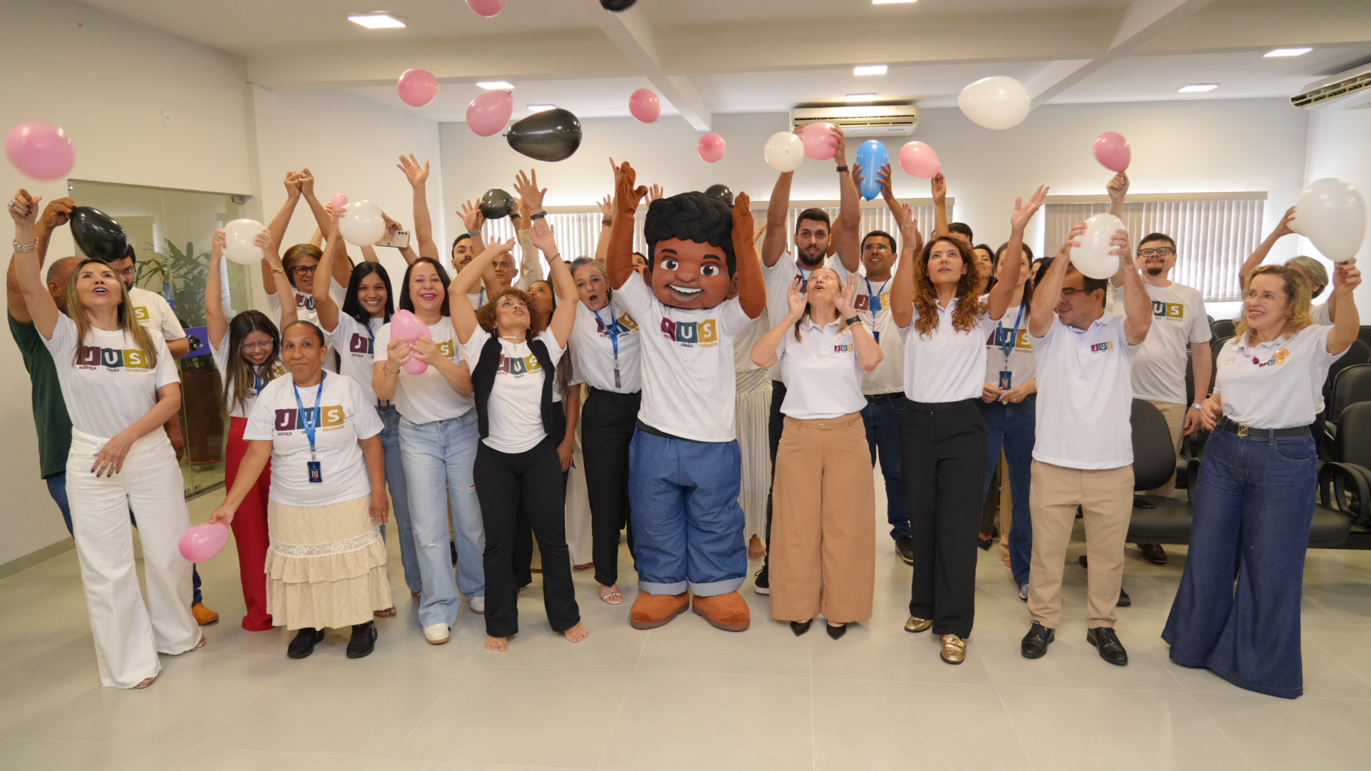 Grupo de pessoas reunidas em ambiente interno celebra com clima descontraído, levantando balões nas cores branca, rosa e preta. Todos vestem camisetas do projeto JUS, e ao centro há um mascote sorridente
