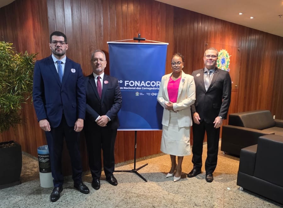 Fotografia em ambiente institucional com quatro pessoas em pé, lado a lado, posando para a câmera. Ao centro, há um banner azul com a inscrição “FONACOR – Fórum Nacional das Corregedorias”. O espaço possui parede de madeira, poltronas pretas e iluminação interna. Os participantes vestem trajes formais: três homens de terno escuro e uma mulher com conjunto claro e blusa rosa. Ao fundo, aparece um brasão na parede, indicando ambiente oficial.