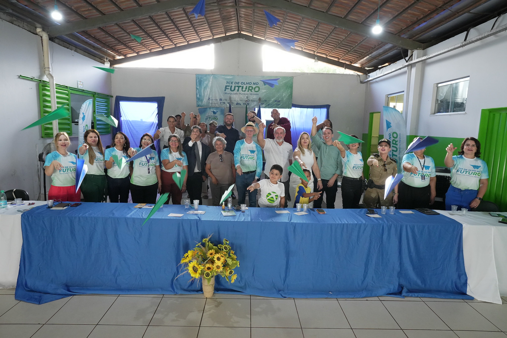 Grupo de participantes posa atrás de uma mesa coberta com toalha azul, em um espaço simples, durante evento do programa “TCE de Olho no Futuro – Aliança pela Primeira Infância”. Ao fundo, há um banner do programa. Em clima descontraído, adultos e duas crianças lançam aviõezinhos de papel coloridos, demonstrando integração e entusiasmo.
