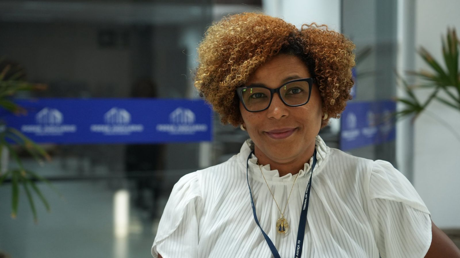 A assistente social Delizângela Alves de Souza posa para foto nas dependências do Fórum de Paraíso do Tocantins após participar da roda de conversa sobre prevenção ao assédio e à discriminação promovida pelo TJTO. Com expressão serena, ela aparece usando blusa branca e crachá institucional, em frente à identidade visual do Poder Judiciário do Tocantins.