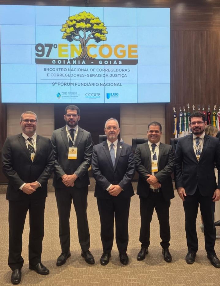 Cinco representantes da Corregedoria-Geral da Justiça do Tocantins posam em pé, usando ternos escuros e crachás, durante o 97º Encoge e o 9º Fórum Fundiário Nacional, em Goiânia (GO). Ao fundo, um painel exibe a identidade visual do evento.