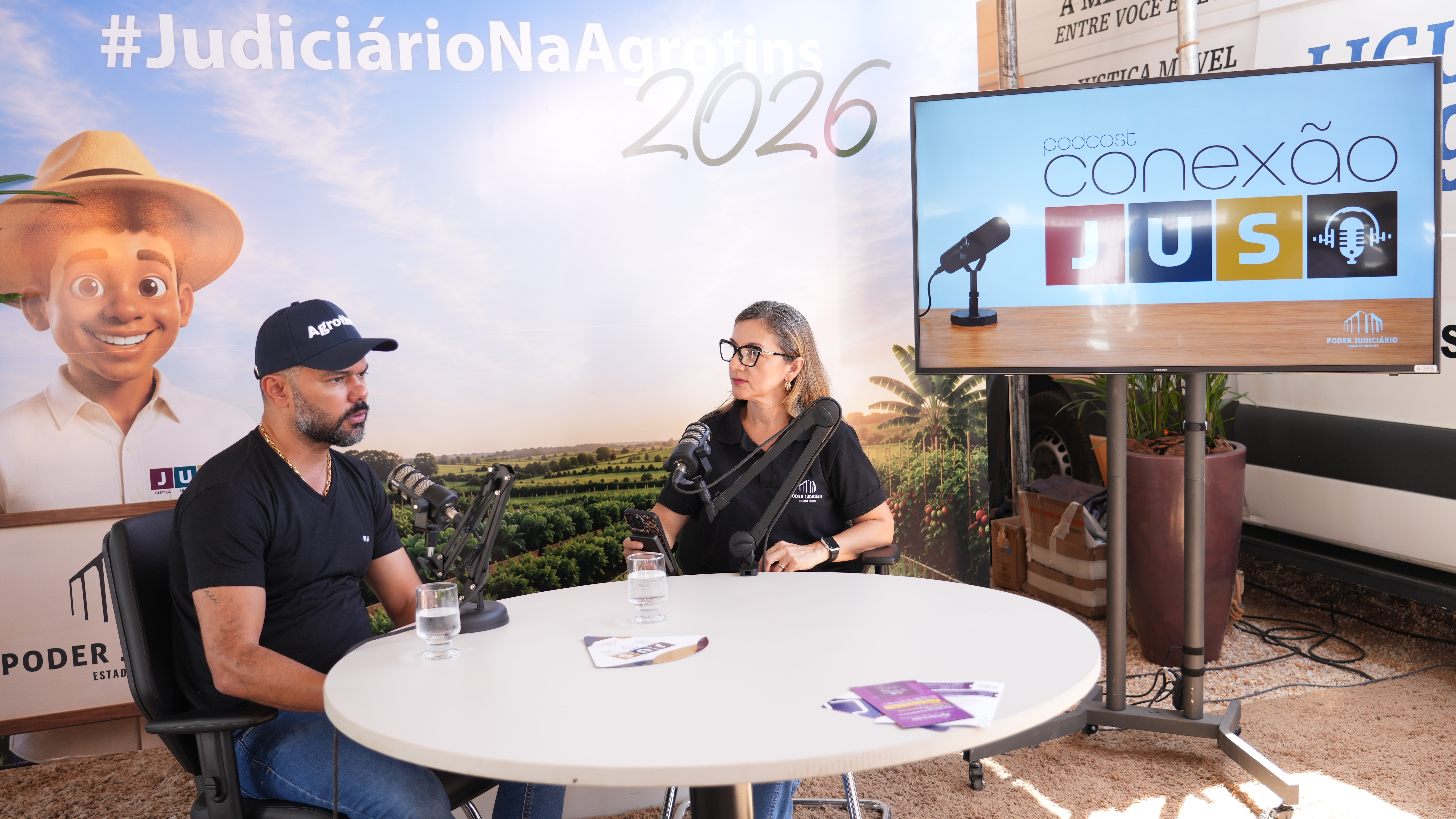 A imagem mostra a gravação do podcast “Conexão JuS” no estande do Poder Judiciário do Tocantins durante a Agrotins 2026. À esquerda, o juiz Wellington Magalhães participa do bate-papo ao lado da jornalista Neuracy Viana. Sobre a mesa, microfones e materiais institucionais compõem o cenário da entrevista, enquanto ao fundo um monitor exibe a identidade visual do podcast e o painel temático da participação do Judiciário na feira.