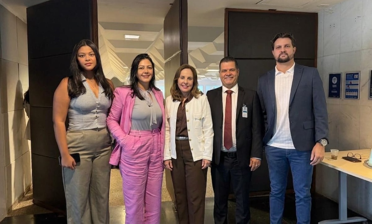 Cinco pessoas posam para foto em ambiente interno do Tribunal de Justiça do Tocantins. Ao centro, uma mulher veste blazer branco e calça marrom. Ao lado, um homem de terno escuro com gravata vermelha e outro homem de blazer azul e camisa branca. À esquerda, duas mulheres, uma com conjunto rosa e outra com blusa cinza e calça bege. O grupo está em pé, próximo à entrada de um auditório ou sala institucional, em momento de encontro institucional.