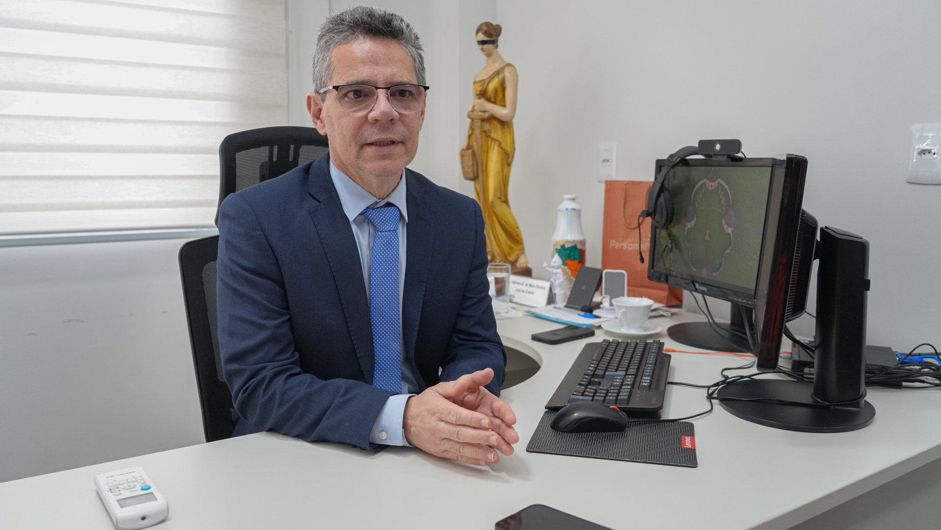 O juiz Adriano Gomes aparece sentado em seu gabinete, durante entrevista ou atendimento institucional. Ele usa terno azul-marinho, camisa clara e gravata azul, e está posicionado diante da mesa de trabalho, com as mãos apoiadas à frente do corpo. Ao lado, há um computador, teclado e itens de escritório. Ao fundo, destaca-se uma escultura da deusa da Justiça.