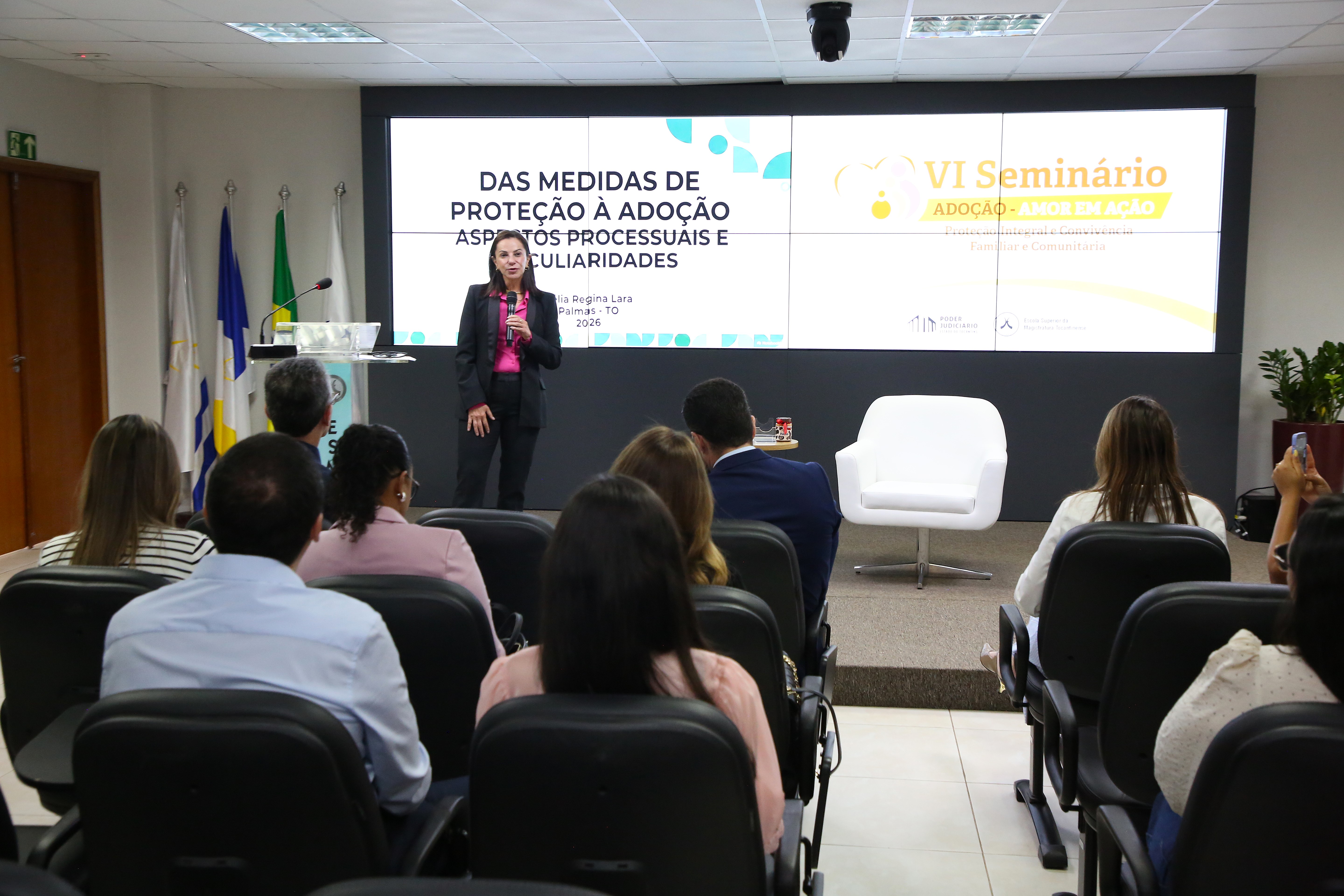 A imagem mostra uma palestrante apresentando o tema “Das Medidas de Proteção à Adoção” durante o VI Seminário Adoção – Amor em Ação, no auditório da Escola Superior da Magistratura Tocantinense.  Enquanto a palestrante fala à frente do público, várias pessoas permanecem sentadas e atentas à apresentação.