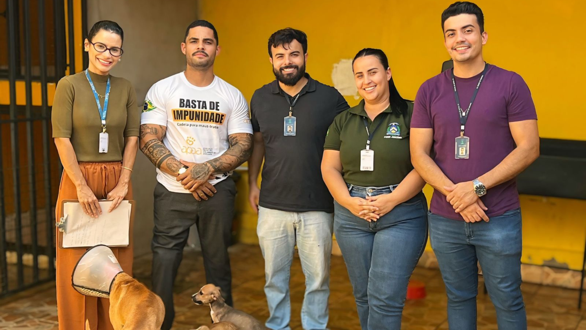 A imagem mostra cinco pessoas posando sorridentes em frente a um prédio, acompanhadas de dois cães, sendo um deles com colar elizabetano.