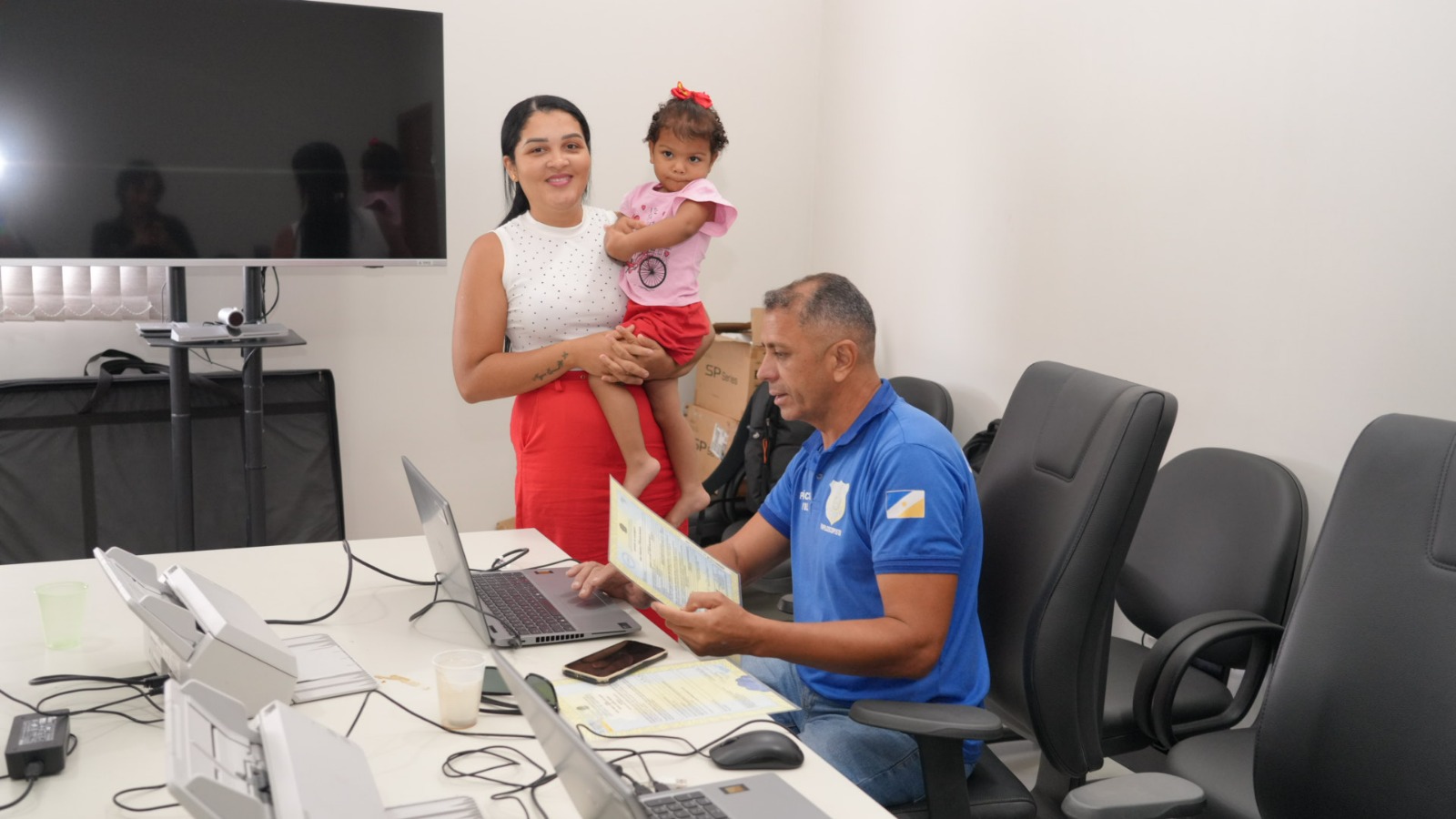 Uma mulher em pé segura uma criança nos braços  ao lado da mesa onde espera atendimento por um servidor público vestido de azul