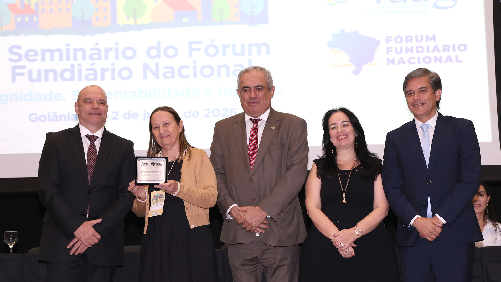 A presidente do Tribunal de Justiça do Tocantins (TJTO), desembargadora Maysa Vendramini Rosal, recebe homenagem durante o Seminário do Fórum Fundiário Nacional, realizado em Goiânia (GO). Na imagem, a magistrada exibe a placa de reconhecimento ao lado de autoridades do sistema de Justiça e representantes de instituições ligadas à temática fundiária. Ao fundo, o painel do evento destaca o tema voltado à dignidade, sustentabilidade e inclusão nas questões fundiárias.
