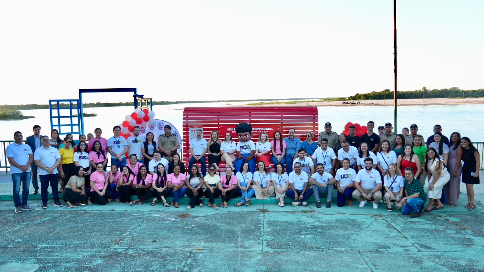 A imagem mostra magistrados, servidores, parceiros e integrantes da comunidade reunidos em frente ao Banco Vermelho, inaugurado em Araguacema durante a programação do projeto Jus em Ação, em um momento de integração e conscientização pelo enfrentamento da violência contra a mulher.  Ao fundo, a paisagem às margens do rio emoldura a fotografia coletiva, que reúne dezenas de participantes posando em frente ao espaço simbólico da campanha.