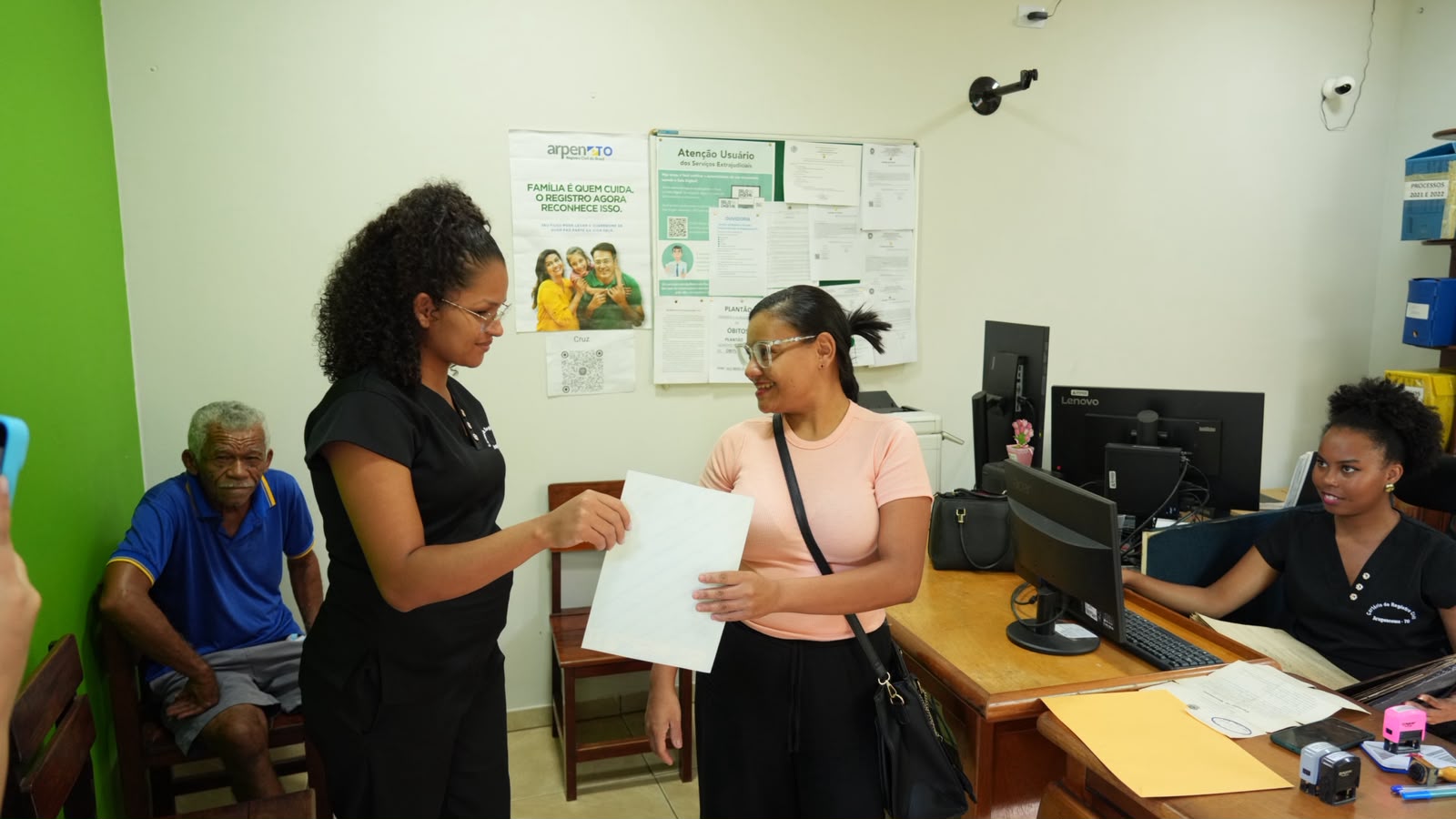 A imagem mostra um atendimento realizado durante o projeto Jus em Ação, com servidoras entregando documentos e orientando a população em um ambiente de serviços. Em destaque, uma cidadã recebe o documento com um sorriso, enquanto outros usuários aguardam atendimento e a equipe realiza os procedimentos administrativos.