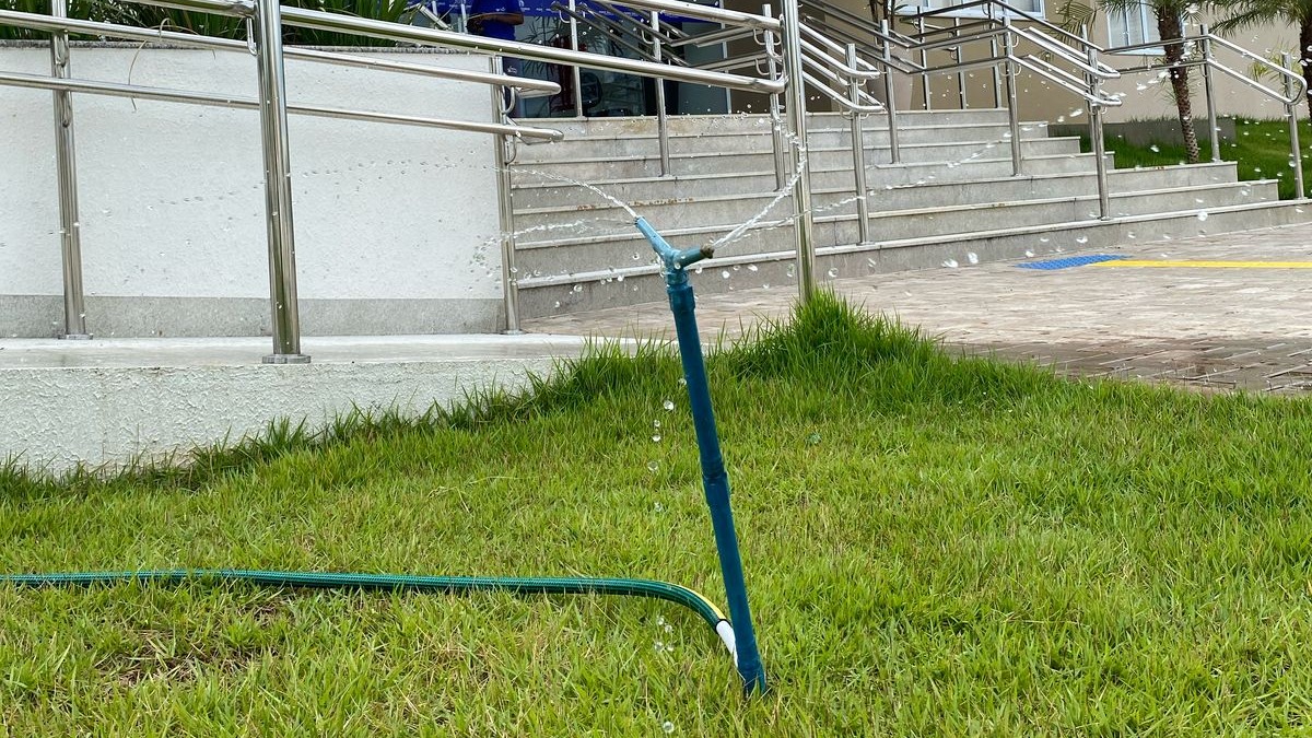A imagem mostra um sistema de irrigação por aspersão instalado em uma área gramada, com um aspersor em funcionamento, lançando jatos de água em diferentes direções para irrigar o jardim. Ao fundo, é possível observar a entrada de um prédio público, com escadaria, piso acessível e corrimãos metálicos, indicando estrutura adequada à acessibilidade.  A cena evidencia o cuidado com a manutenção das áreas verdes e o uso planejado da água para irrigação do paisagismo, contribuindo para a conservação ambiental e a boa apresentação do espaço institucional.