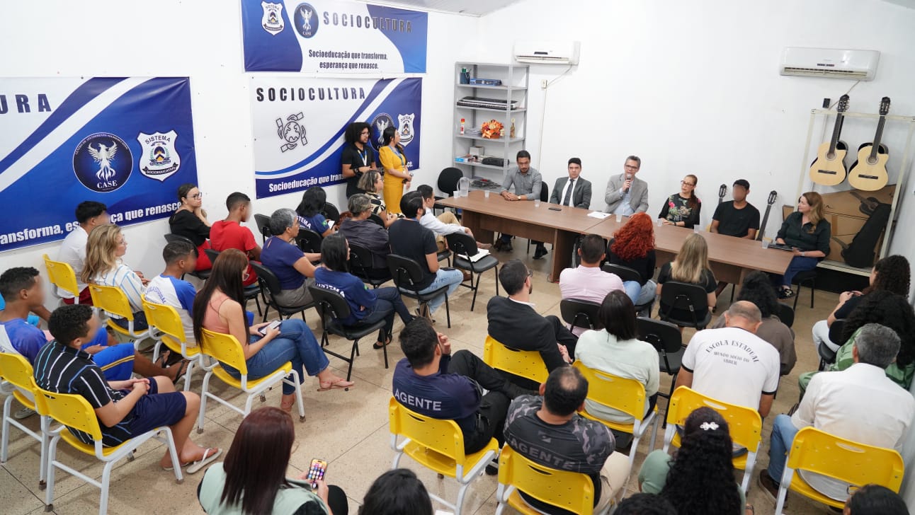 Pessoas sentadas em cadeiras organizadas em auditório acompanham reunião institucional. À frente, uma mesa com autoridades e convidados sentados, sendo um dos participantes em pé falando ao microfone. O ambiente é interno, com banners institucionais ao fundo e instrumentos musicais apoiados na parede lateral.