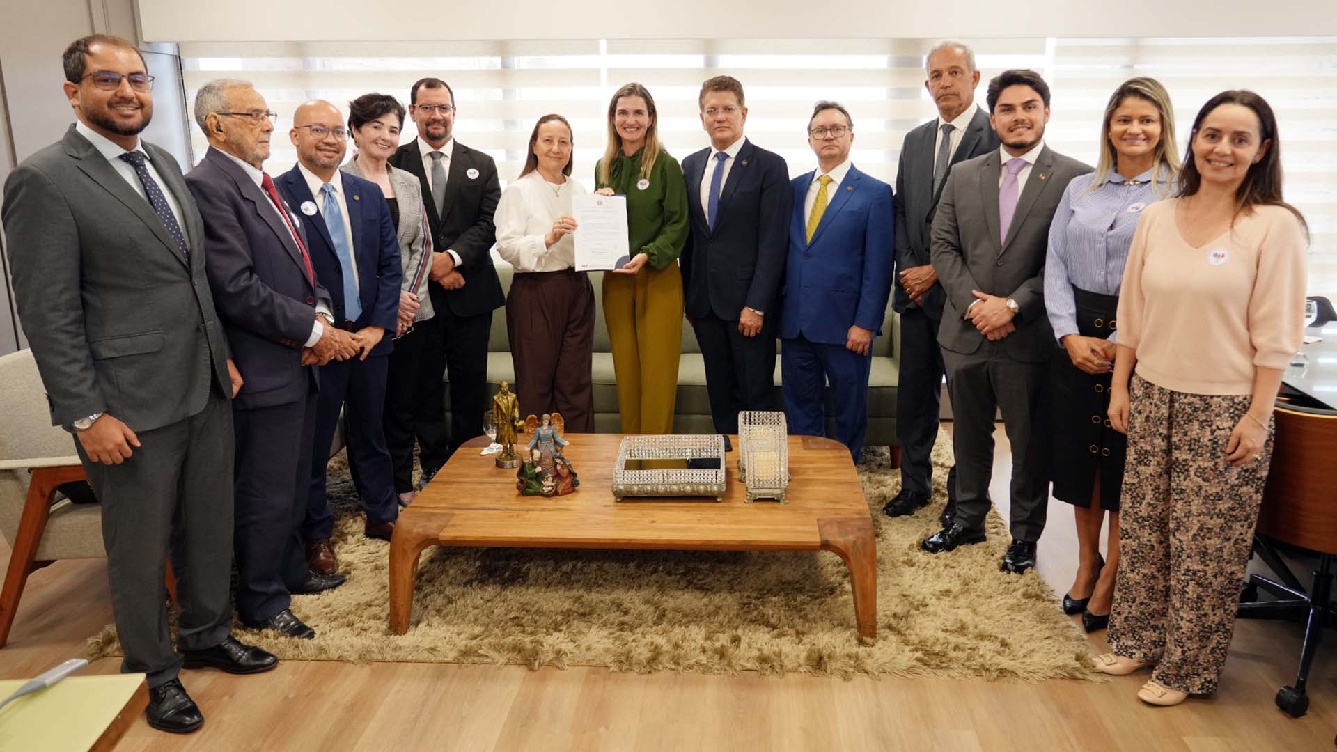 Presidente do TJTO ao centro, desembargadores e advogados em pé posam para foto. A presidente do TJTO e a presidente em exercício da OAB-TO, ao centro, seguram um documento. À frente, mesa de madeira com objetos decorativos sobre tapete. Ao fundo, sofá e persianas claras. Todos vestem trajes formais.
