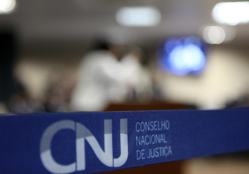 A imagem mostra uma faixa com a sigla CNJ e a identificação do Conselho Nacional de Justiça em primeiro plano. Ao fundo, desfocado, aparece um ambiente institucional.