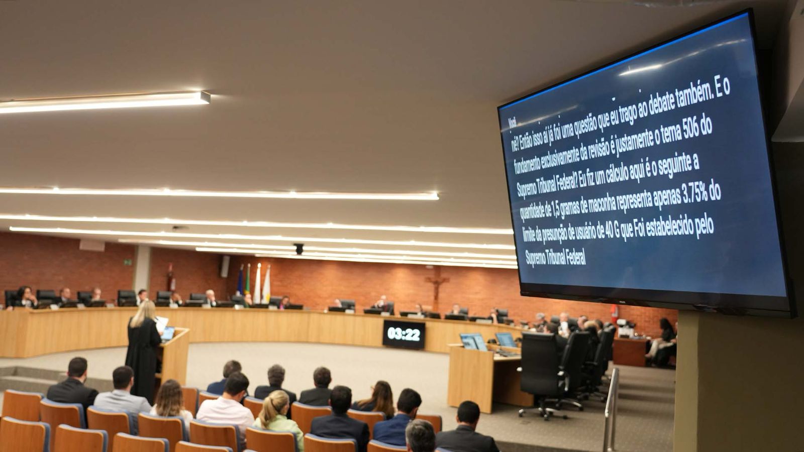 Sessão plenária em auditório, com magistrados sentados à mesa semicircular. Uma pessoa fala ao púlpito. À direita, um telão exibe texto de legenda. Público acompanha a sessão em cadeiras dispostas em fileiras. Ambiente amplo e iluminado.