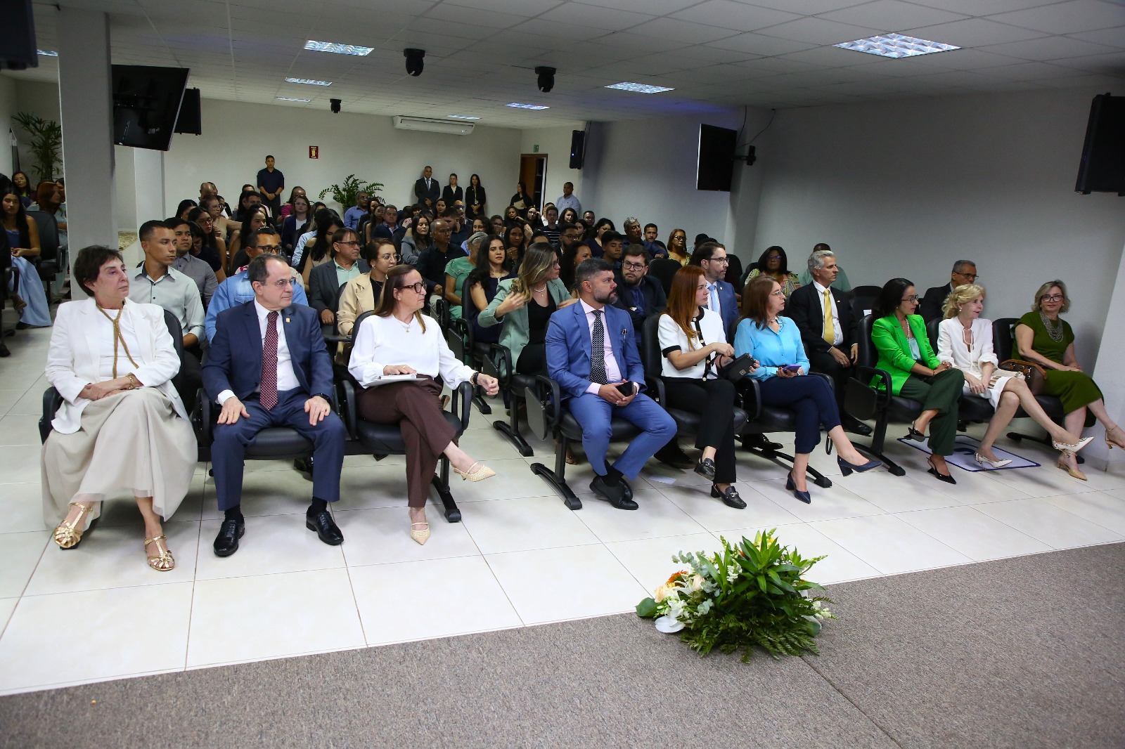 A imagem mostra um auditório cheio, com autoridades e público sentados em cadeiras organizadas, acompanhando a certificação da Turma III do Programa de Residência em Prática Jurídica. O ambiente é interno, formal, com telões laterais, iluminação clara e um arranjo floral à frente do espaço.