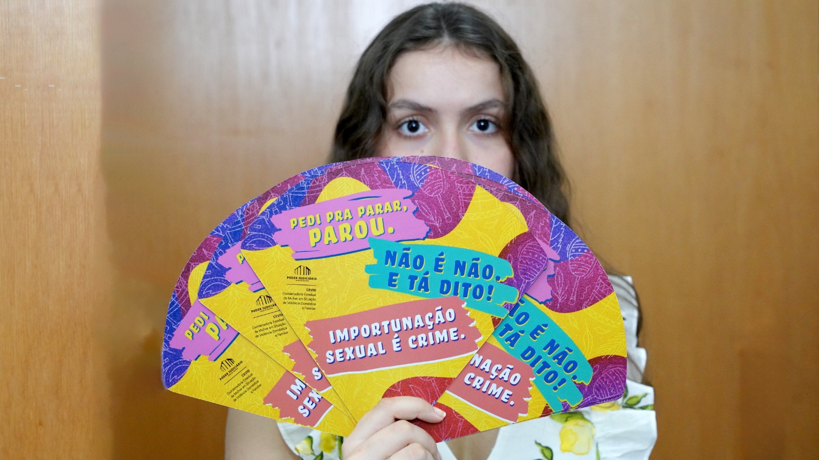 Jovem diante de fundo de madeira segura leques coloridos com as frases “Pedi pra parar, parou.”, “Não é não, e tá dito!” e “Importunação sexual é crime.”  Apenas os olhos ficam visíveis. Nos leques consta a identificação do Poder Judiciário e da Coordenadoria da Mulher em Situação de Violência Doméstica.