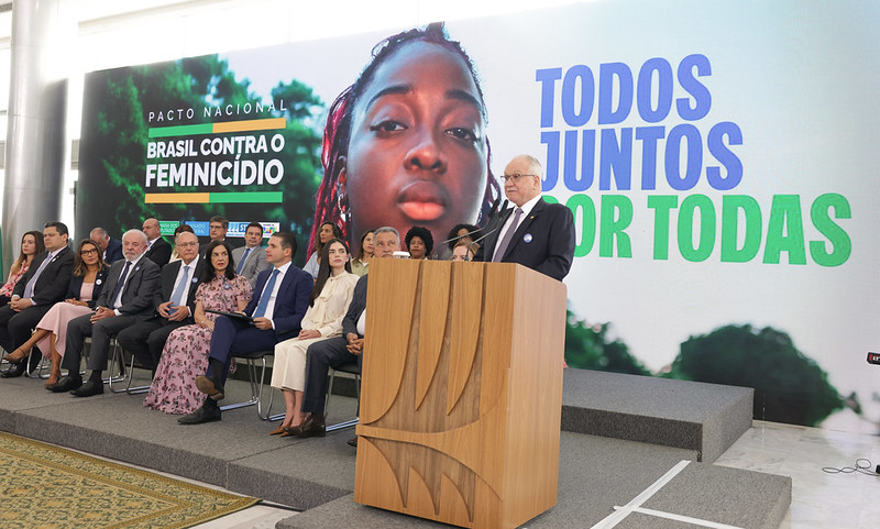 A imagem mostra uma solenidade institucional voltada ao enfrentamento da violência contra a mulher. Em primeiro plano, um homem de terno discursa em um púlpito de madeira. Ao fundo, um grande painel exibe a frase “Pacto Nacional: Brasil contra o Feminicídio” e o slogan “Todos juntos por todas”, ao lado do rosto de uma mulher em destaque