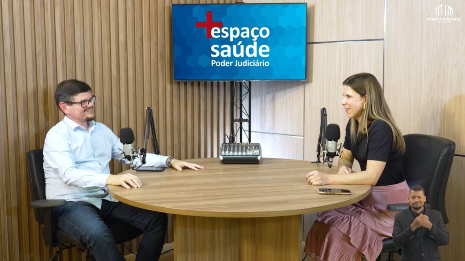 A imagem mostra um estúdio do Poder Judiciário, com duas pessoas sentadas frente a frente, em uma mesa redonda, durante a gravação do PodCast para conscientização no Dia Nacional de Combate às Drogas e ao Alcoolismo. Ao fundo, aparece o painel “+ Espaço Saúde”.