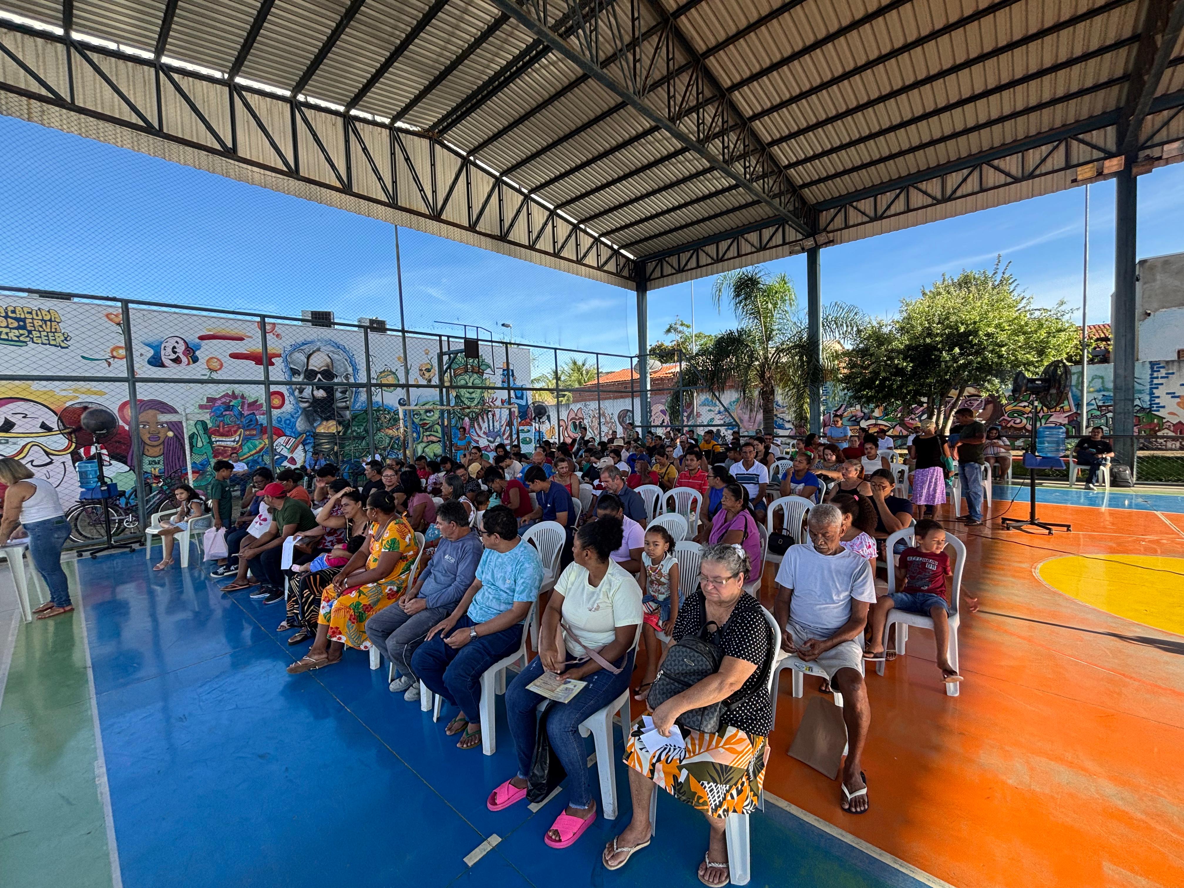 A imagem mostra uma quadra poliesportiva coberta, com o chão pintado em azul e laranja. No local, dezenas de pessoas — entre adultos, idosos e crianças — estão sentadas em cadeiras plásticas brancas, organizadas em fileiras, como em um evento comunitário ou ação social.  Ao fundo, há um muro com grafites coloridos e artísticos. A quadra é cercada por grades e tem estrutura metálica no teto.