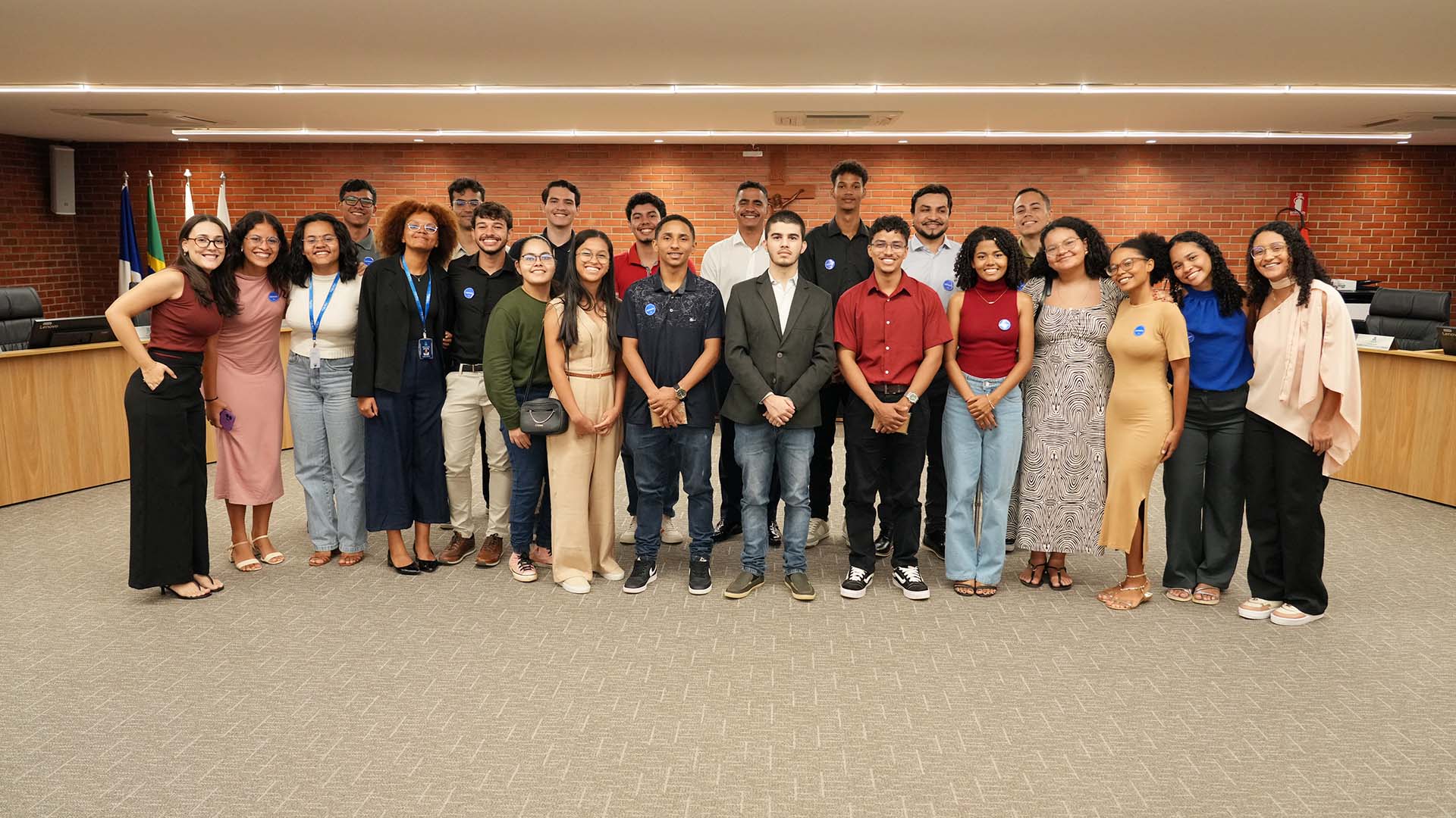 Grupo de jovens estudantes posa para foto no salão do Tribunal Pleno, com parede de tijolos ao fundo e bandeiras à esquerda. Estão organizados em duas fileiras, em frente a uma mesa de plenário.