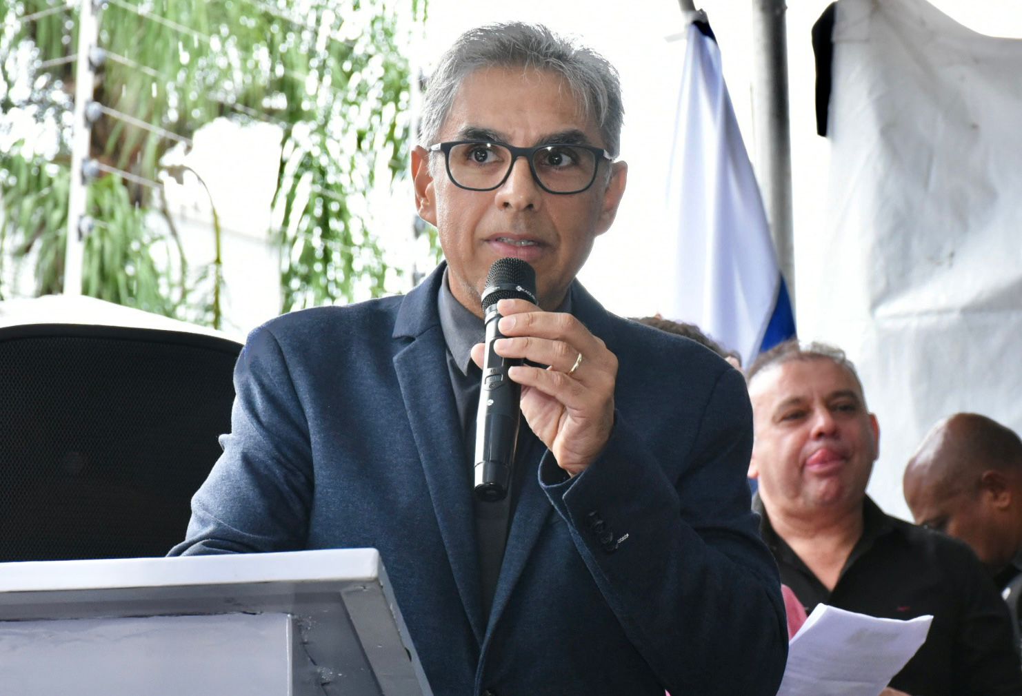 De terno azul-escuro e camisa preta, o juiz Jossanner Nery segura um microfone e fala ao público. Usa óculos de armação escura e tem cabelos grisalhos. A imagem foi registrada ao ar livre, com estrutura predial ao fundo e árvores à direita. 