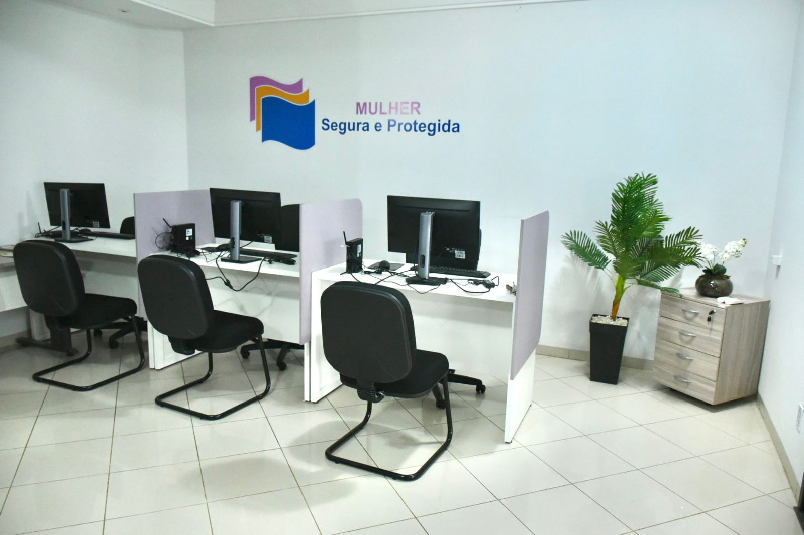 Sala com três estações de atendimento equipadas com computadores e cadeiras pretas. Na parede, há a identificação “Mulher Segura e Protegida”. O ambiente é claro, organizado e conta com planta decorativa e pequeno armário lateral.