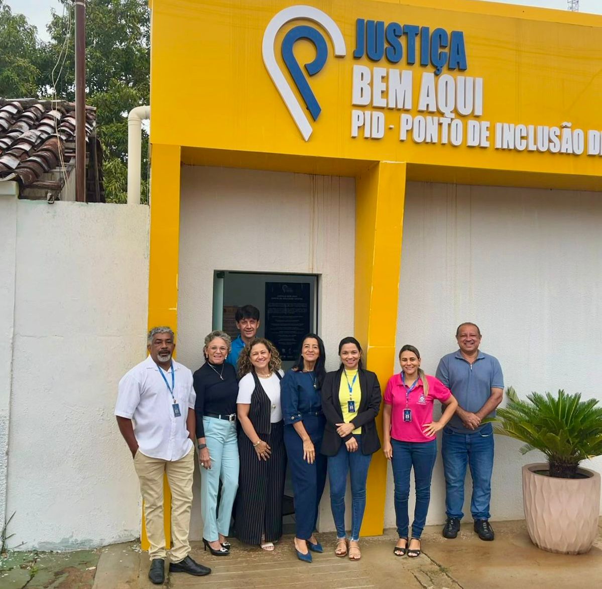 Equipe do Judiciário posa em frente à unidade do “Justiça Bem Aqui – PID (Ponto de Inclusão Digital)”, em Almas. O grupo está reunido na entrada do espaço, identificado por fachada amarela com a logomarca do programa. Ao fundo, a porta de acesso à unidade e, ao lado, um vaso com planta ornamental compõem o cenário da foto institucional.