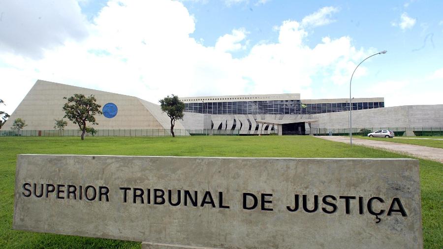 A imagem mostra a sede do Superior Tribunal de Justiça (STJ), em Brasília, com sua arquitetura moderna e ampla área gramada à frente. Em primeiro plano, aparece uma placa de concreto com a inscrição “Superior Tribunal de Justiça”, diante do edifício principal ao fundo.
