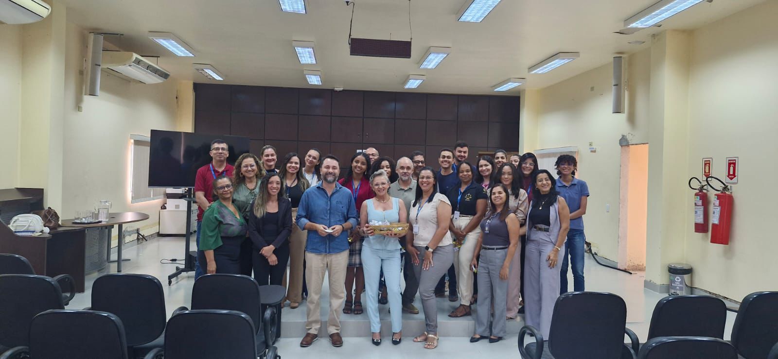 Grupo de magistrados(as), servidores(as) e integrantes da equipe do NAPsi posa para foto no auditório da Comarca de Dianópolis. Ao centro, participantes seguram uma lembrança simbólica, enquanto os demais se organizam em pé, sorrindo, em clima de integração. À frente, cadeiras do plenário aparecem vazias, e ao fundo é possível ver painel de madeira, televisão e estrutura de som do espaço.