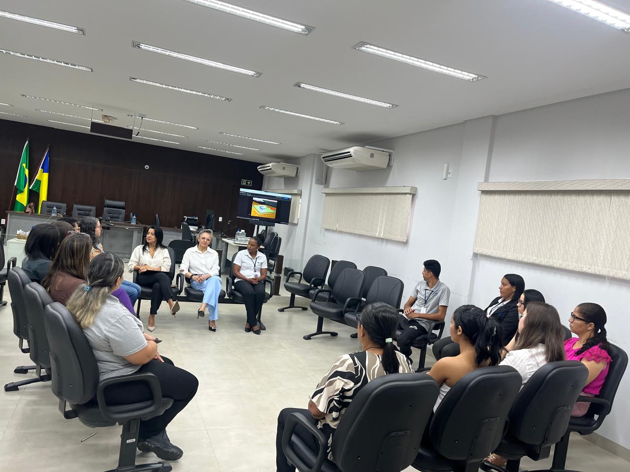 Servidores(as) participam de roda de conversa conduzida pela equipe do NAPsi em sala de audiência da Comarca de Taguatinga. Sentados(as) em círculo, os(as) participantes acompanham atentamente as facilitadoras, em ambiente organizado com cadeiras acolchoadas, painel de madeira ao fundo e bandeiras oficiais posicionadas ao lado da bancada.