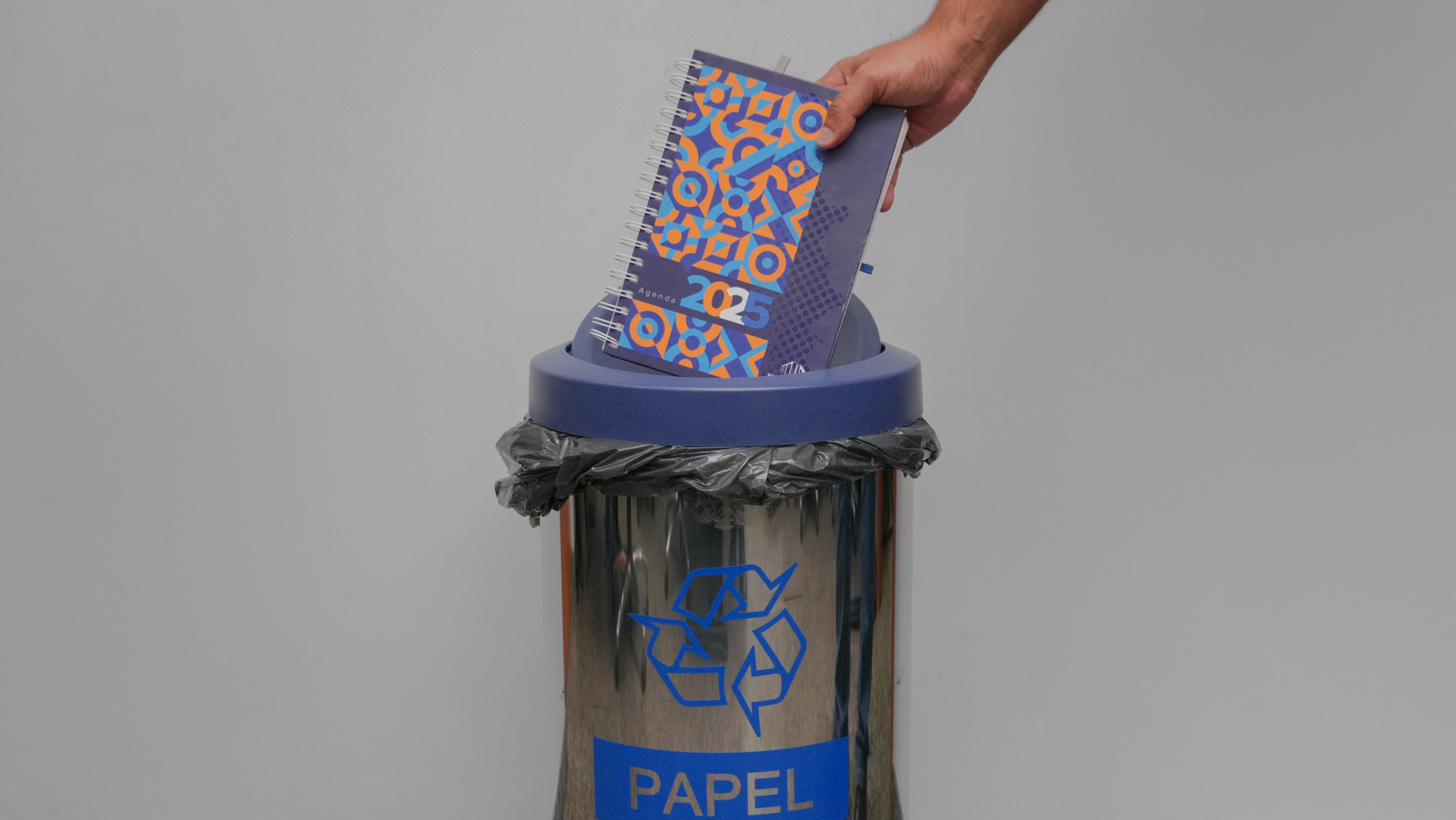 A imagem mostra uma ação de descarte consciente de papel. Em primeiro plano, uma mão deposita uma agenda do ano de 2025 em um coletor metálico de reciclagem identificado com o símbolo universal da reciclagem e a inscrição “PAPEL”, em destaque na cor azul. O coletor possui aro superior também azul e saco plástico interno para acondicionamento dos materiais descartados. A agenda tem capa espiralada, com design gráfico colorido em tons de azul e laranja, evidenciando que se trata de material impresso já fora de uso. O fundo neutro e claro reforça o foco na prática sustentável e na correta destinação de resíduos recicláveis.