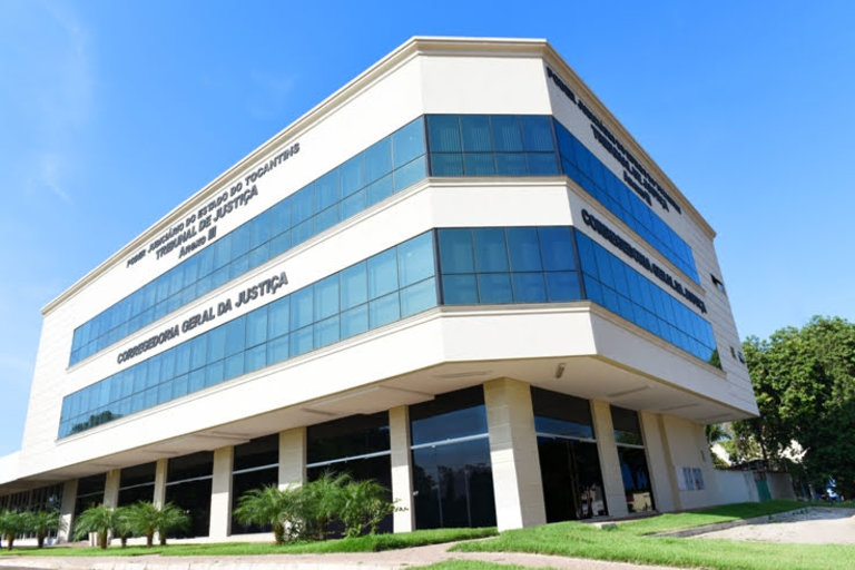 A imagem mostra a fachada da Corregedoria-Geral da Justiça do Tocantins, com estrutura em concreto e amplas áreas de vidro espelhado. O prédio está inserido em um ambiente arborizado, com jardins na área externa e céu azul ao fundo.