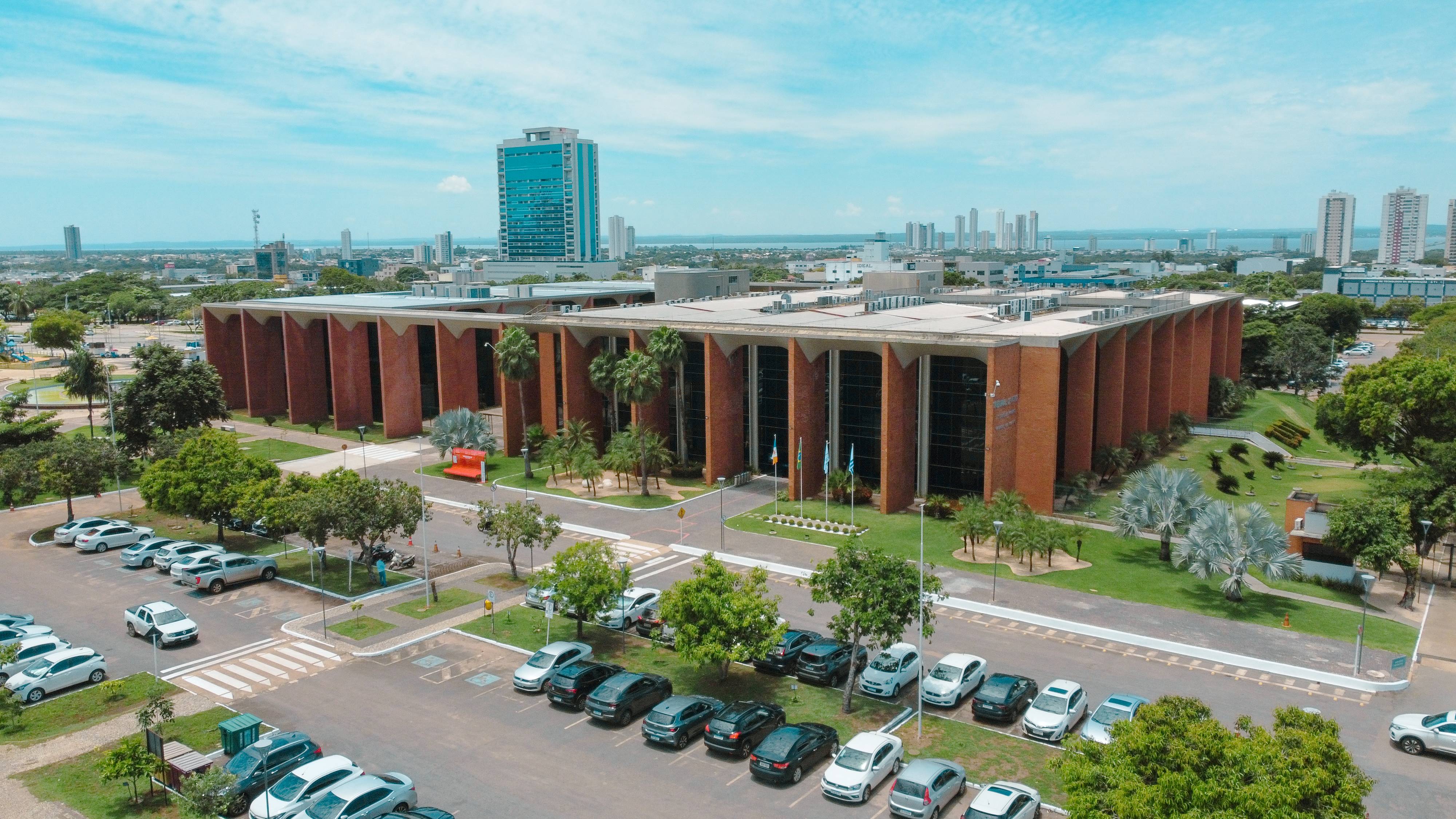A imagem mostra o prédio institucional do Tribunal de Justiça do Tocantins, com arquitetura imponente em tijolos aparentes, amplas áreas envidraçadas e paisagismo bem cuidado ao redor. Ao fundo, vê-se a área urbana com prédios altos, vias organizadas e estacionamento.