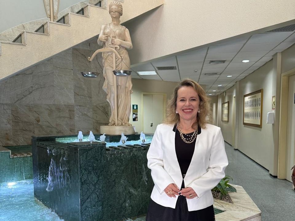 A imagem mostra a juíza Cirlene de Assis em pé, sorridente, vestida com blazer branco e saia preta, em um ambiente institucional interno. Ao fundo, há uma fonte com escultura da deusa da Justiça, escadas curvas e um corredor bem iluminado.