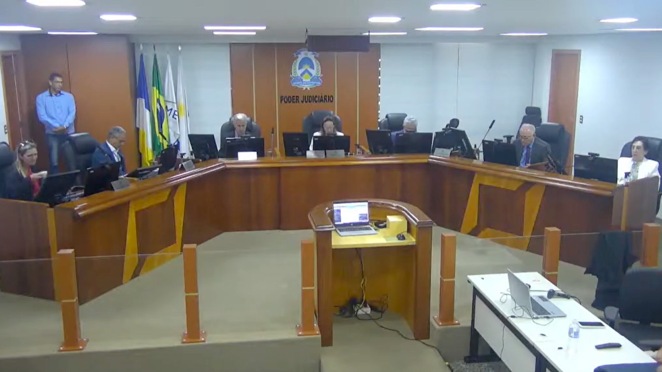 Imagem aberta de uma sessão no TJTO; na foto aparece uma mesa grande de madeira com desembargadoes e demais autoridades sentadas; ao fundo o brasão da Justiça na parede e bandeiras