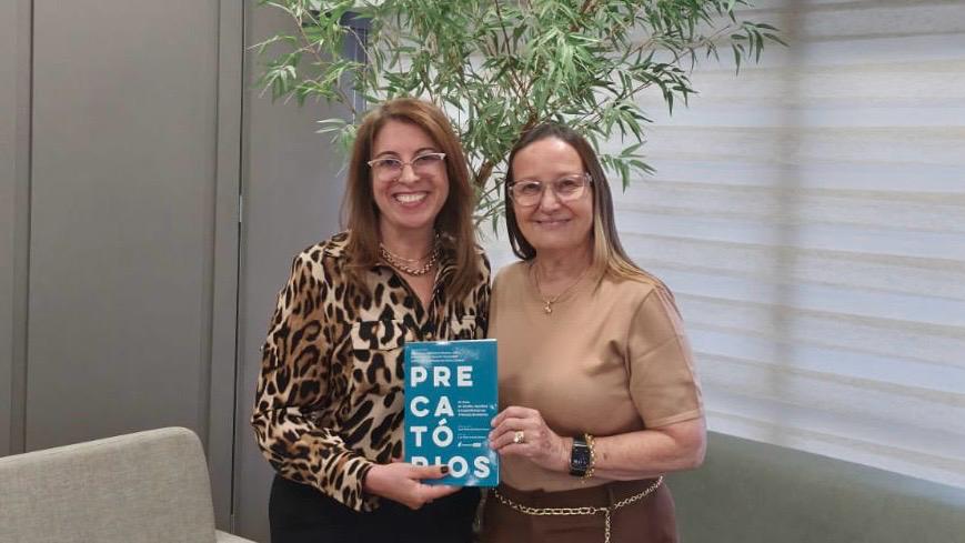 A imagem mostra duas mulheres posam lado a lado em um ambiente interno, com aparência institucional e iluminação suave. Ambas sorriem para a câmera, transmitindo cordialidade e formalidade. A mulher à esquerda veste uma blusa de estampa animal print em tons de marrom e preto, usa óculos de armação clara e segura um livro de capa azul com o título “Precatórios” em destaque. A mulher à direita usa blusa de tom bege, também com óculos, e apoia o livro junto à colega, indicando um momento de entrega ou apresentação da obra.
