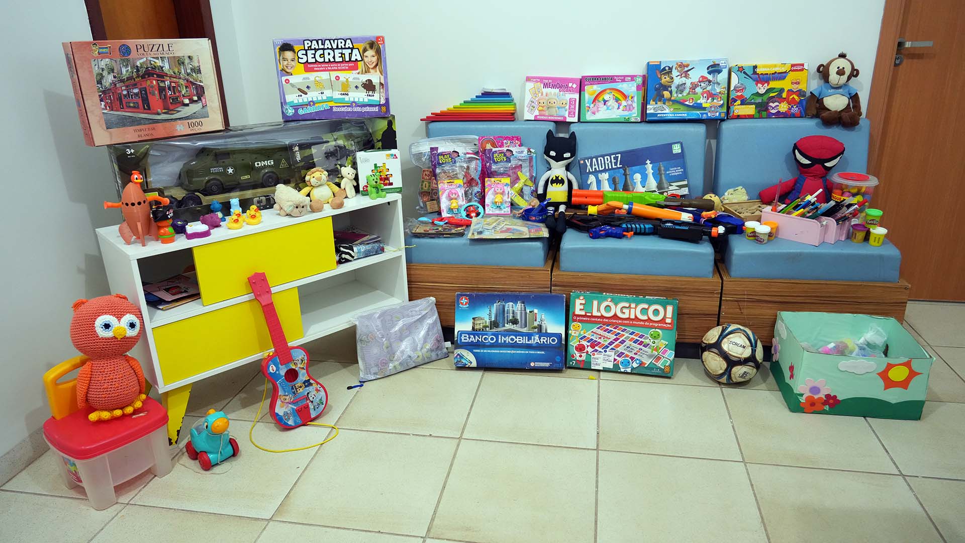 A imagem mostra um espaço organizado com diversos brinquedos e materiais lúdicos infantis dispostos sobre um banco estofado azul, uma pequena estante branca com gavetas amarelas e no chão. Entre os itens expostos há jogos educativos, como quebra-cabeça, jogo da memória, xadrez, Banco Imobiliário e jogos de lógica, além de brinquedos variados, como bonecos de super-heróis, pelúcias, carrinhos, pistolas de brinquedo, massinhas de modelar e instrumentos musicais infantis, como um pequeno violão.  Também aparecem materiais de desenho e pintura, guardados em um organizador rosa, e objetos artesanais, como uma coruja de crochê sobre um banquinho infantil. No chão há ainda uma bola de futebol, um carrinho e caixas organizadoras com mais brinquedos.